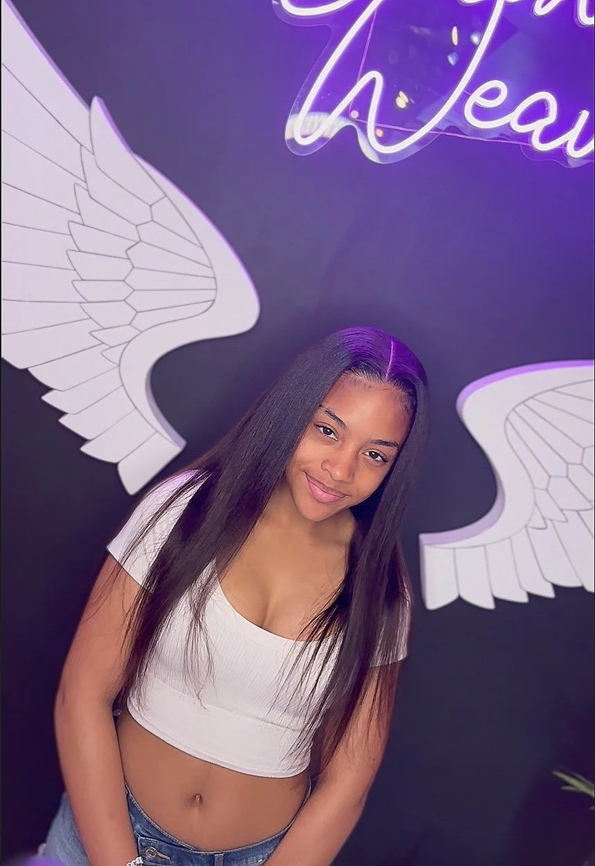 A young woman with long straight black hair wearing a white crop top and blue jeans, standing in front of a dark wall with neon purple lighting, angel wings, and a neon sign.