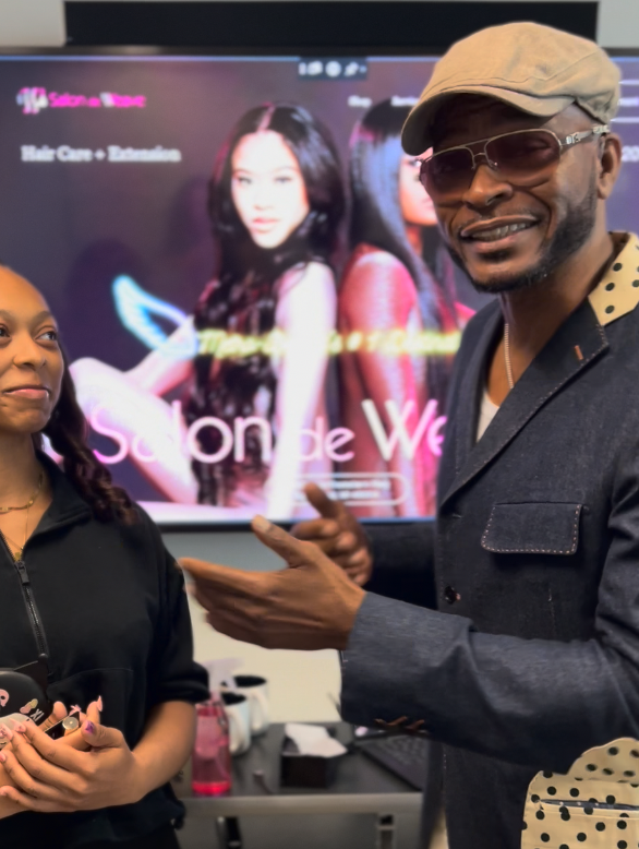 A man wearing sunglasses, a beige cap, and a dark blazer with polka dot lining on the collar, is speaking and gesturing with his hands to a young woman with braids, dressed in black. They are standing in front of a screen showing a promotional image of two women and text that reads 'Salon de We' and 'Hair Care + Education.'