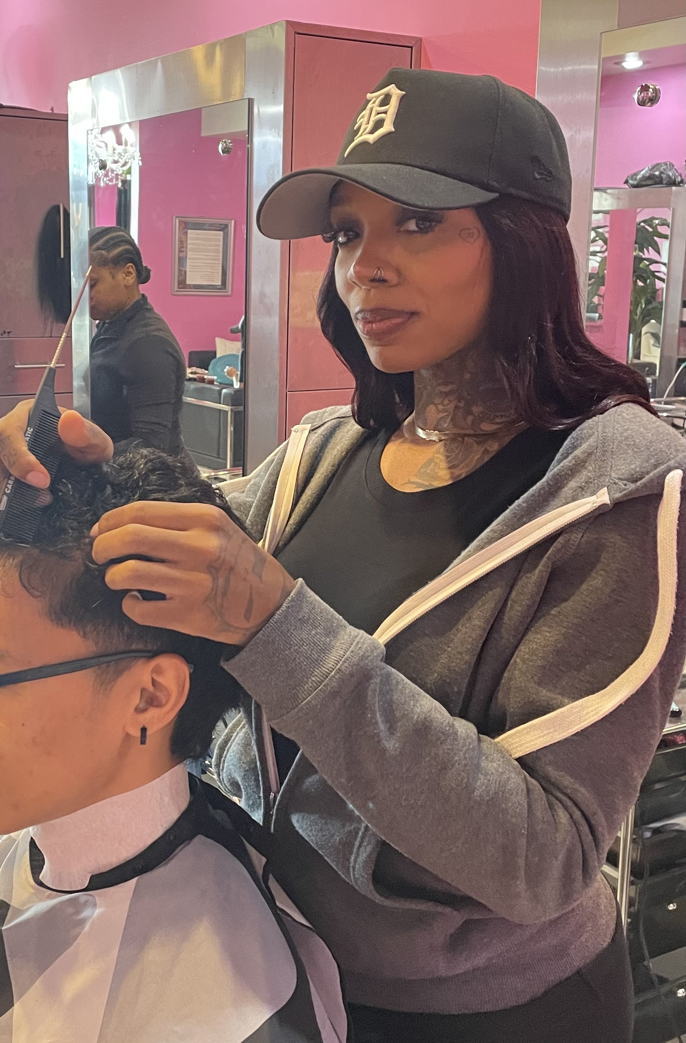 A woman with tattoos and a Detroit Tigers cap is giving a haircut to a man in a salon with pink walls and mirrors.