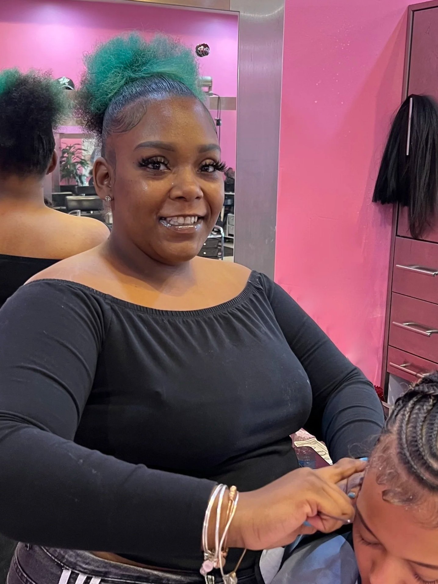 A woman with green hair styled in a high puff, wearing a black off-the-shoulder top, is smiling at the camera inside a salon with pink walls. She appears to be getting her hair styled or braided by another person.