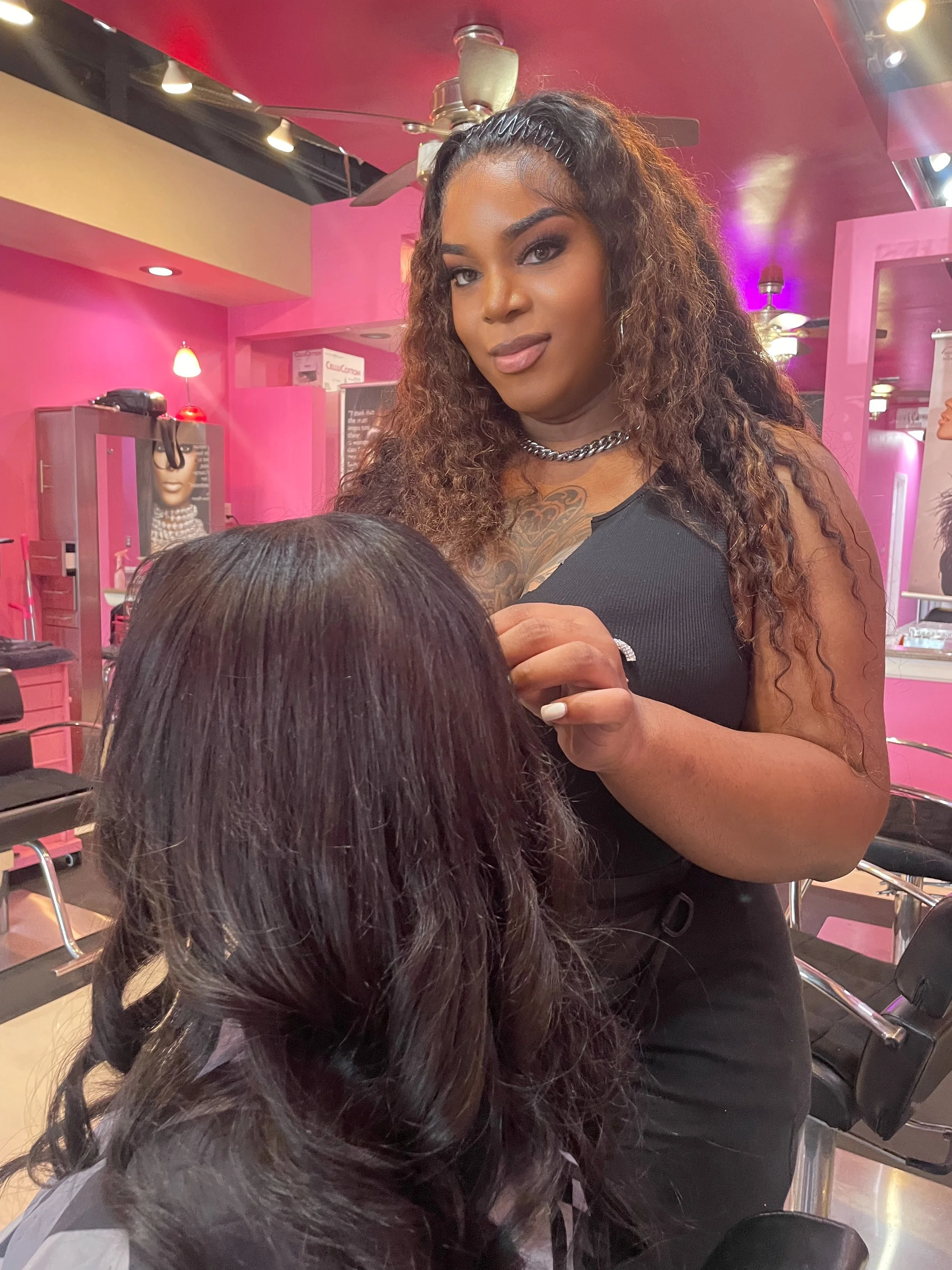 A woman with long, curly hair and makeup, wearing a black top and chain necklace, is standing in a pink-themed salon or beauty shop, working on a client's hair.