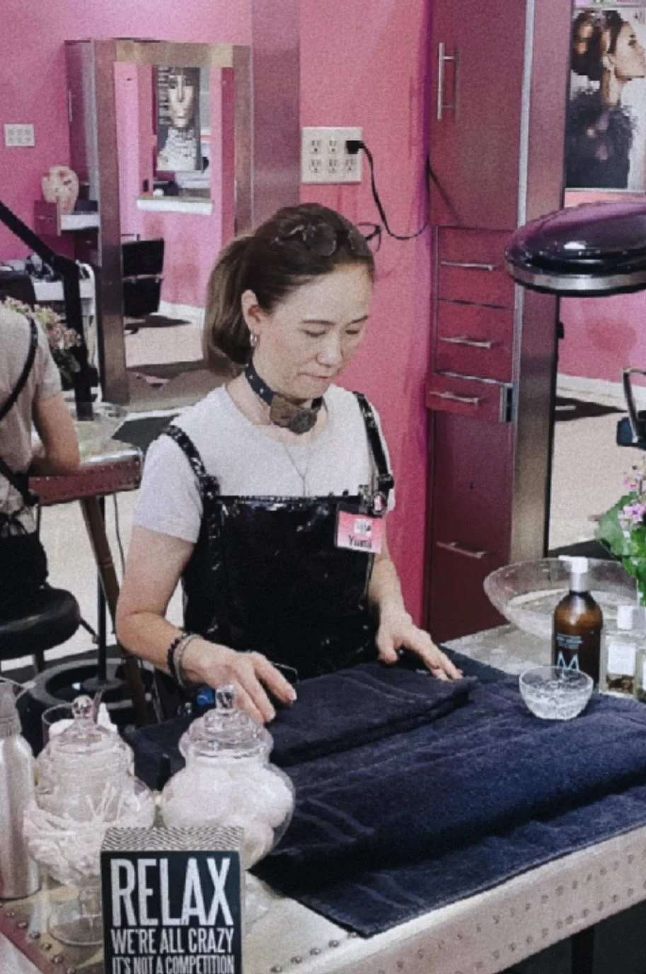 A woman in a salon or boutique setting with a pink wall, a mirror, and salon furniture. She is wearing a black apron over a white shirt and a black choker, and is standing at a table with various decor items and bottles, including a sign that says "RELAX."