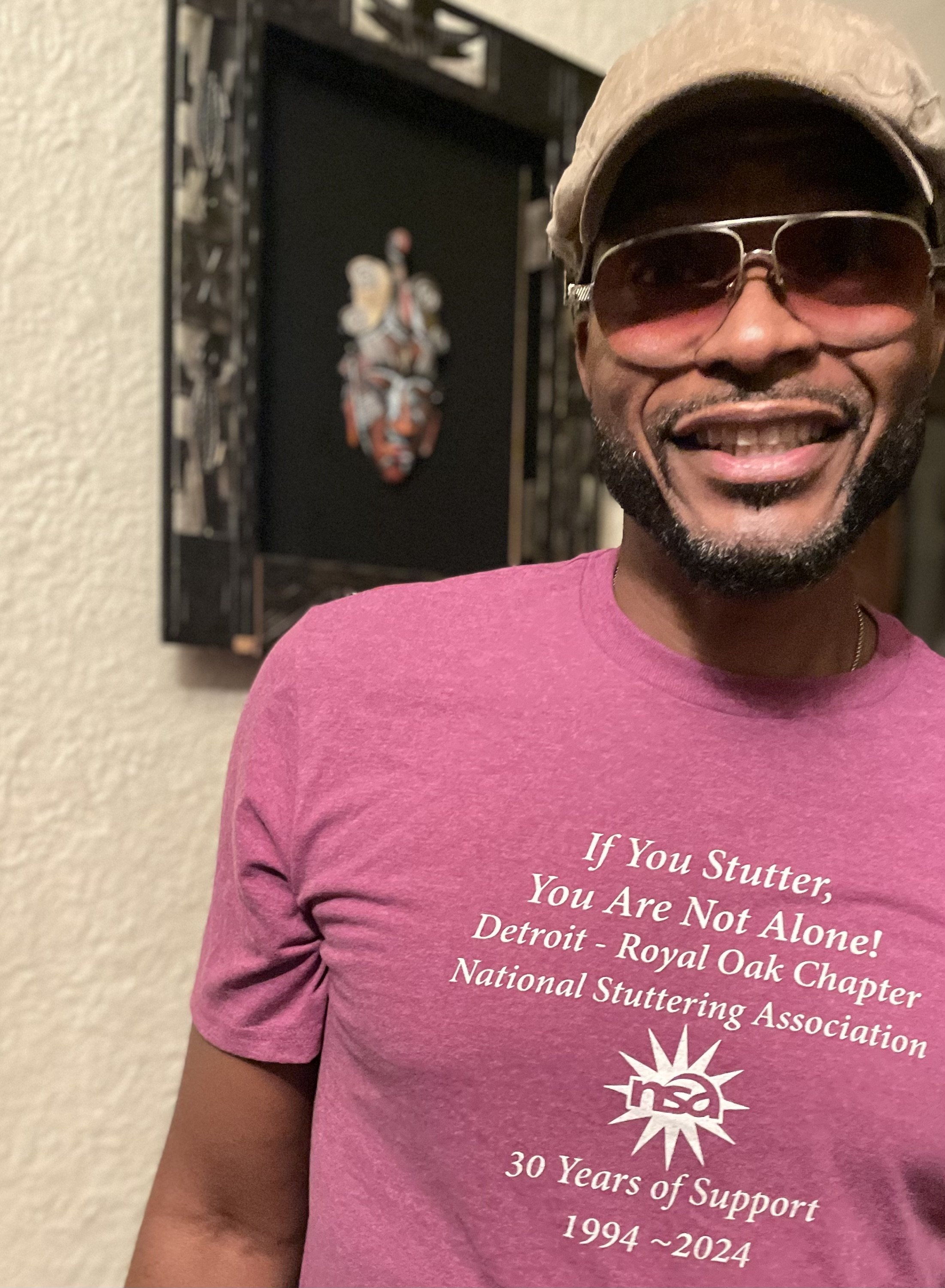 A person wearing sunglasses, a cap, and a pink shirt with white text celebrating 30 years of support for the Detroit Royal Oak Chapter of the National Stuttering Association, from 1994 to 2024, is smiling in front of a black decorative wall.