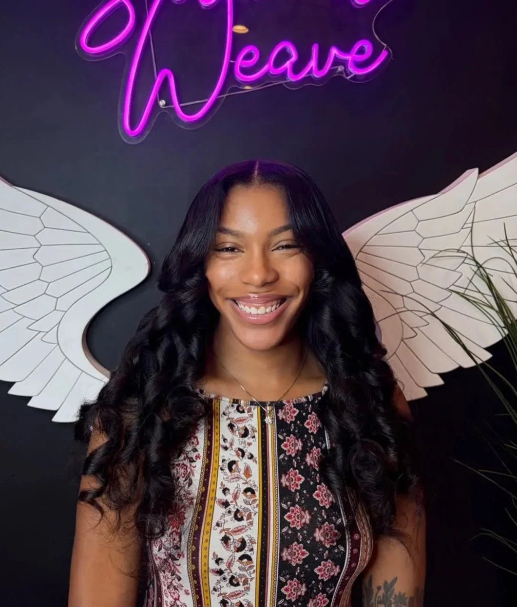 A smiling woman with long, curly black hair, wearing a sleeveless patterned dress, standing in front of a black background with white angel wings and a neon purple sign that says 'Leave'.