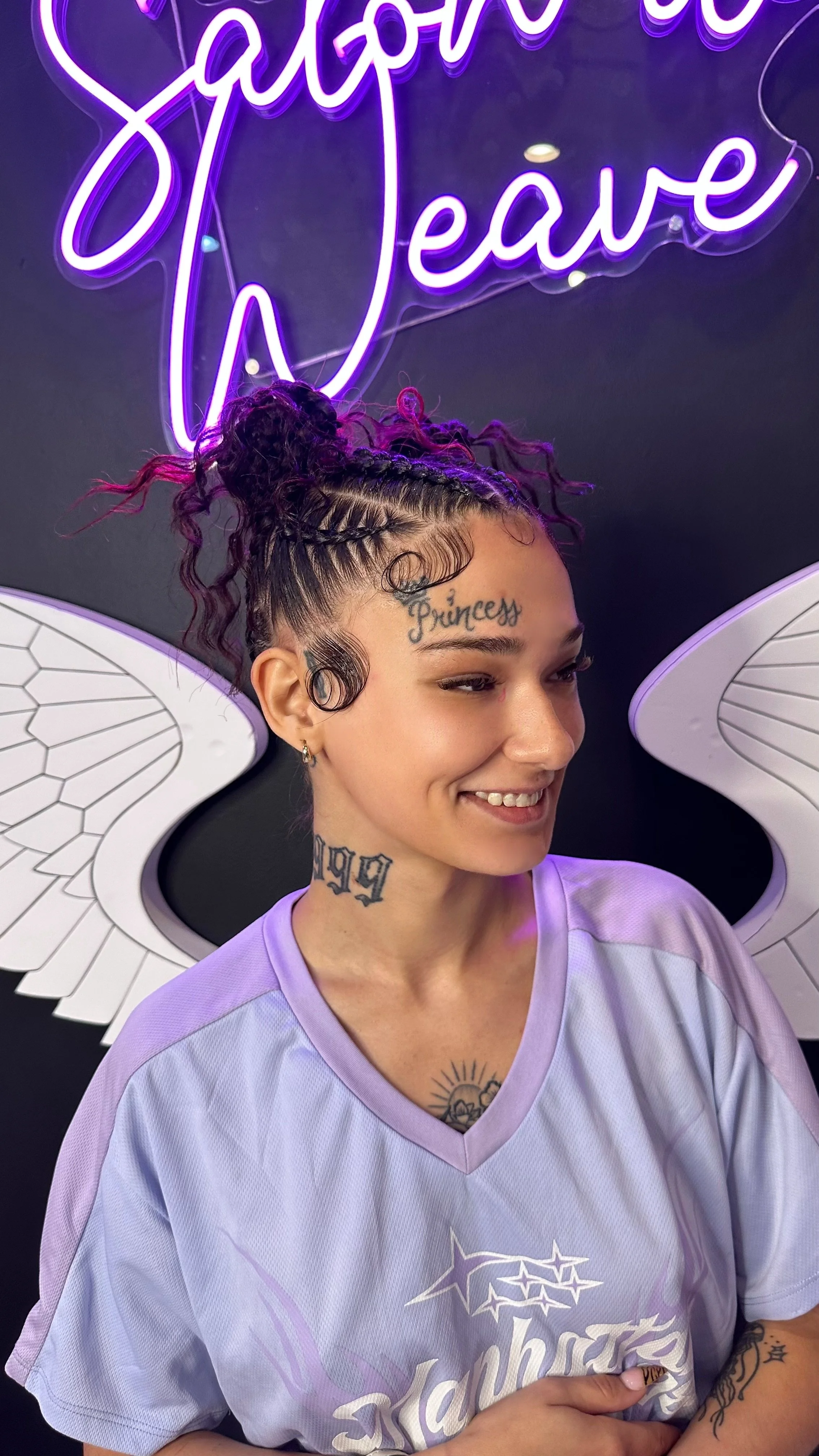 A woman with purple and black curly hair styled in a bun, wearing a light purple sports jersey and angel wings, smiling in front of a neon sign reading 'Sapphire Leave' with purple and white lighting.