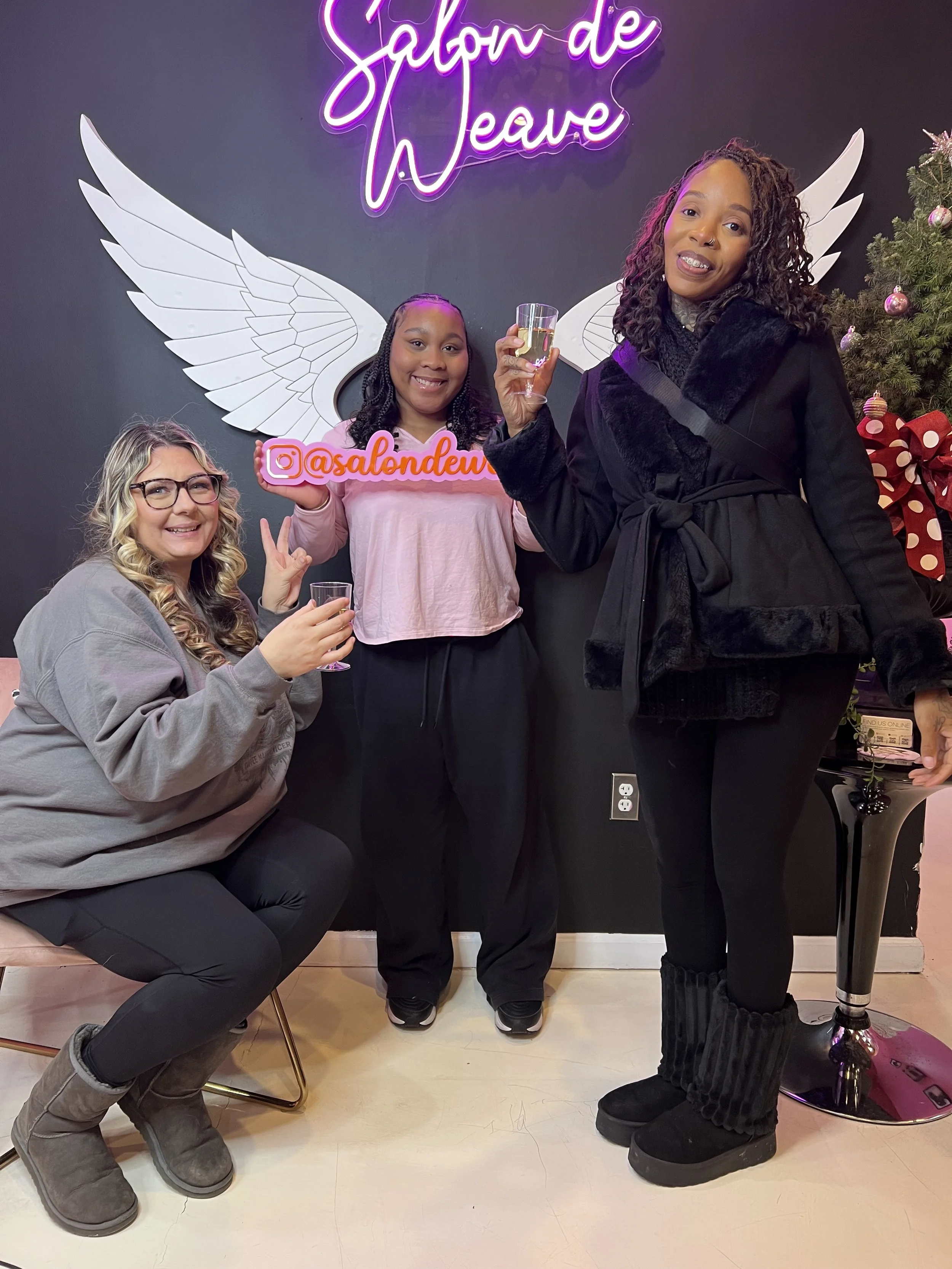 Three women posing indoors in front of a wall with a neon sign that says 'Salon de Weave', white angel wings decoration, and a Christmas tree. The woman on the right is holding a glass of champagne, and the woman in the center is holding an Instagram sign that says '@asalendew'.