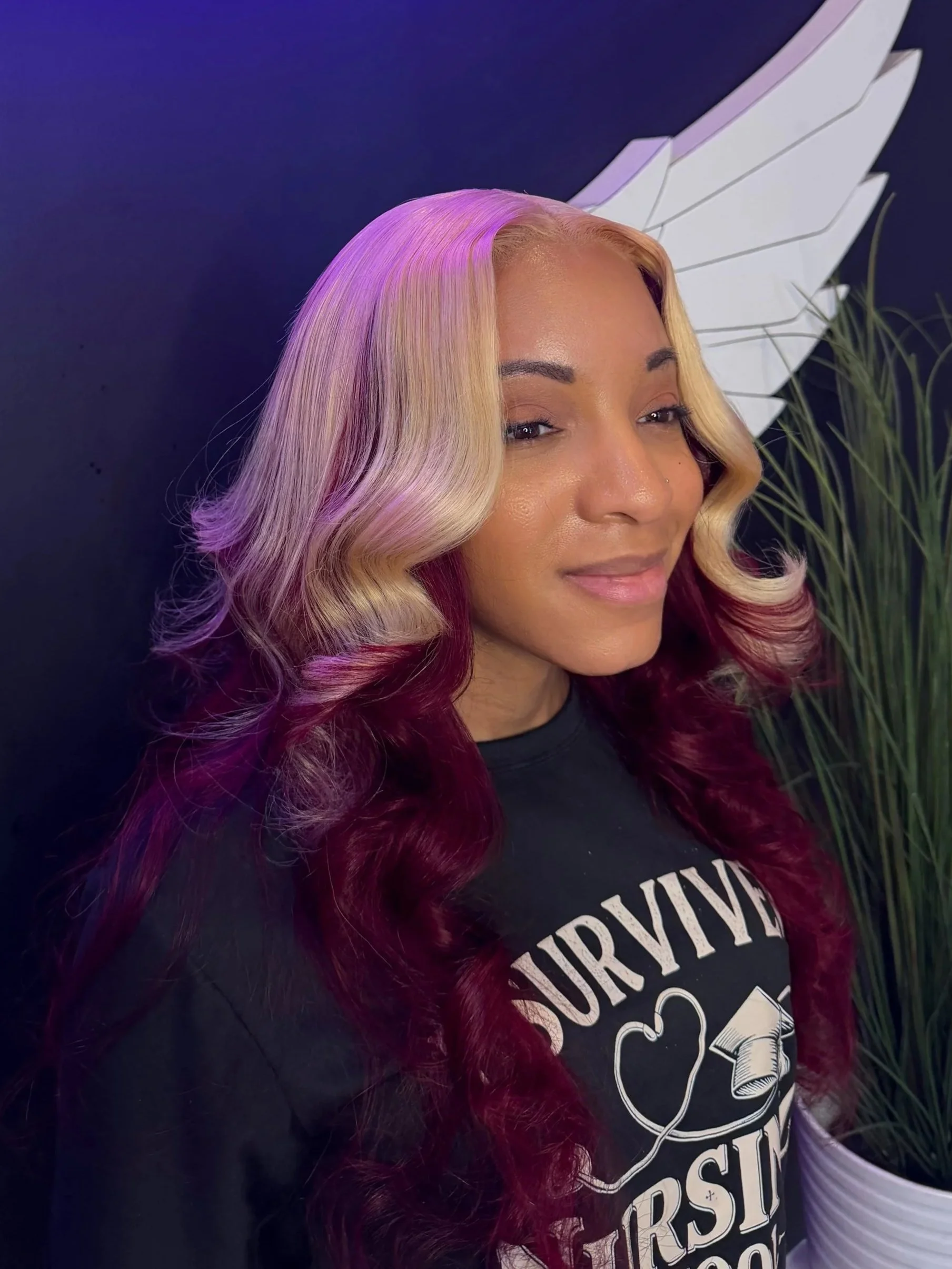 Close-up of a woman with long wavy hair dyed in blonde and burgundy, smiling slightly, standing in front of a black background with white decorative wings on the wall.