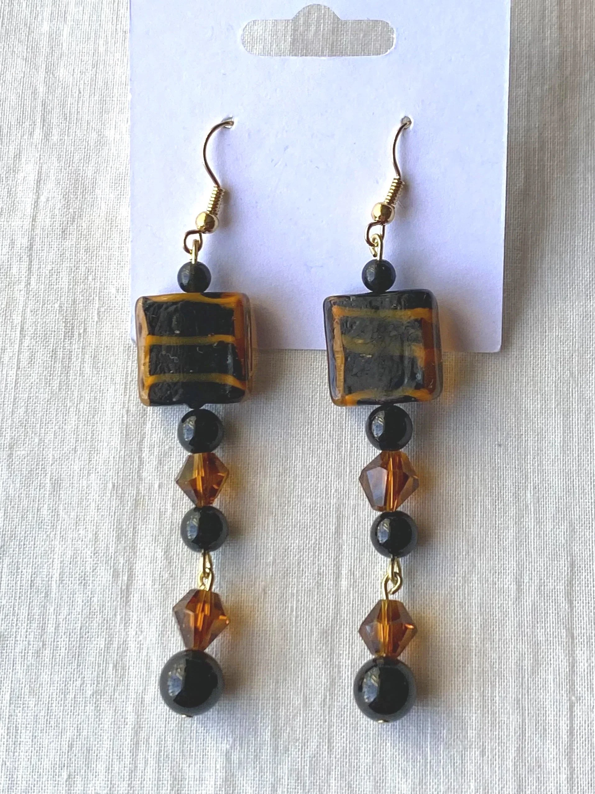 A pair of earrings featuring square-shaped beads with a black and amber swirling pattern, accented with small black and amber beads, hanging from metal hooks.
