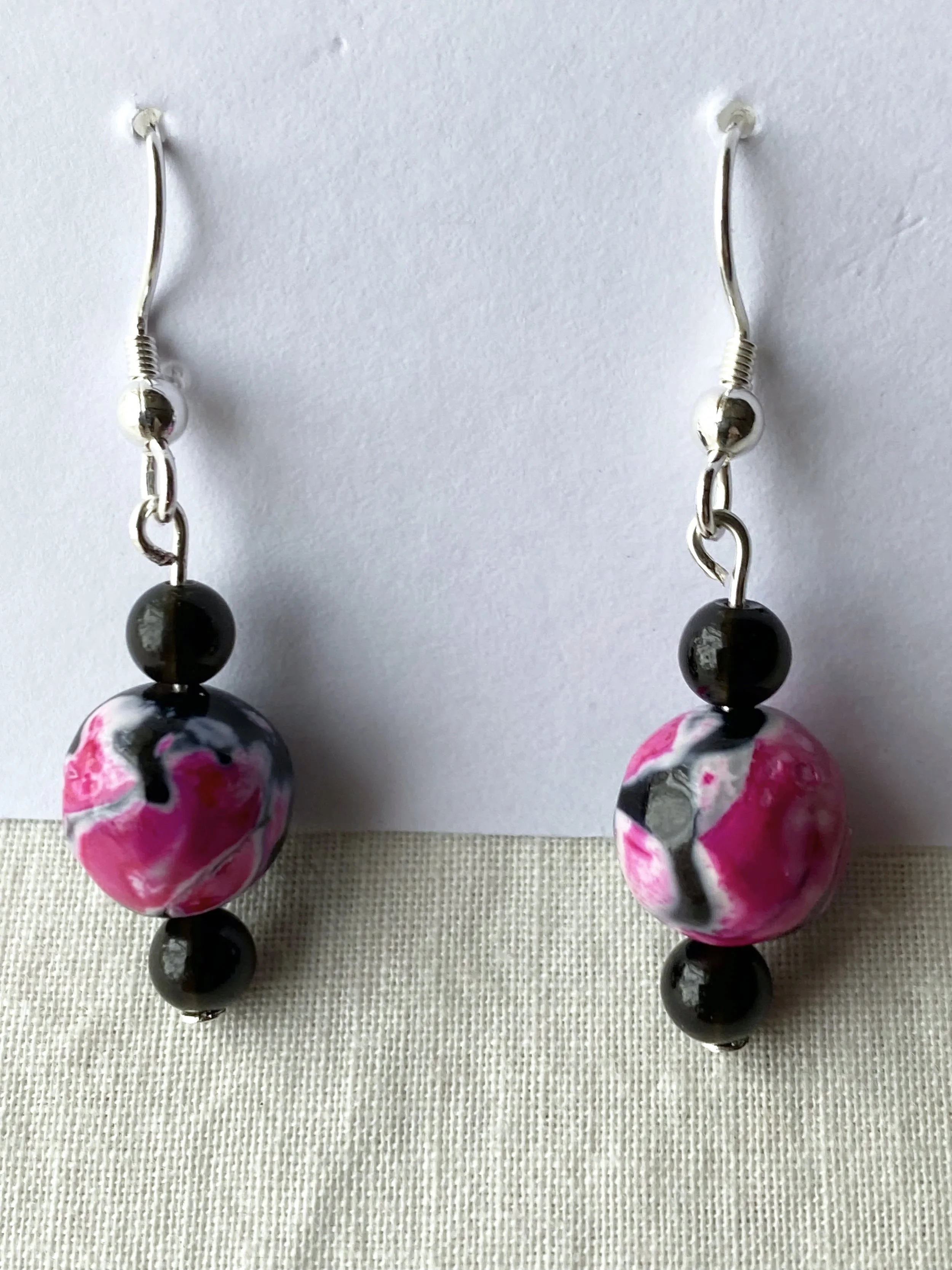 A pair of earrings with large marble-like pink and black beads, smaller black beads, and metal hooks, hanging from a white background.