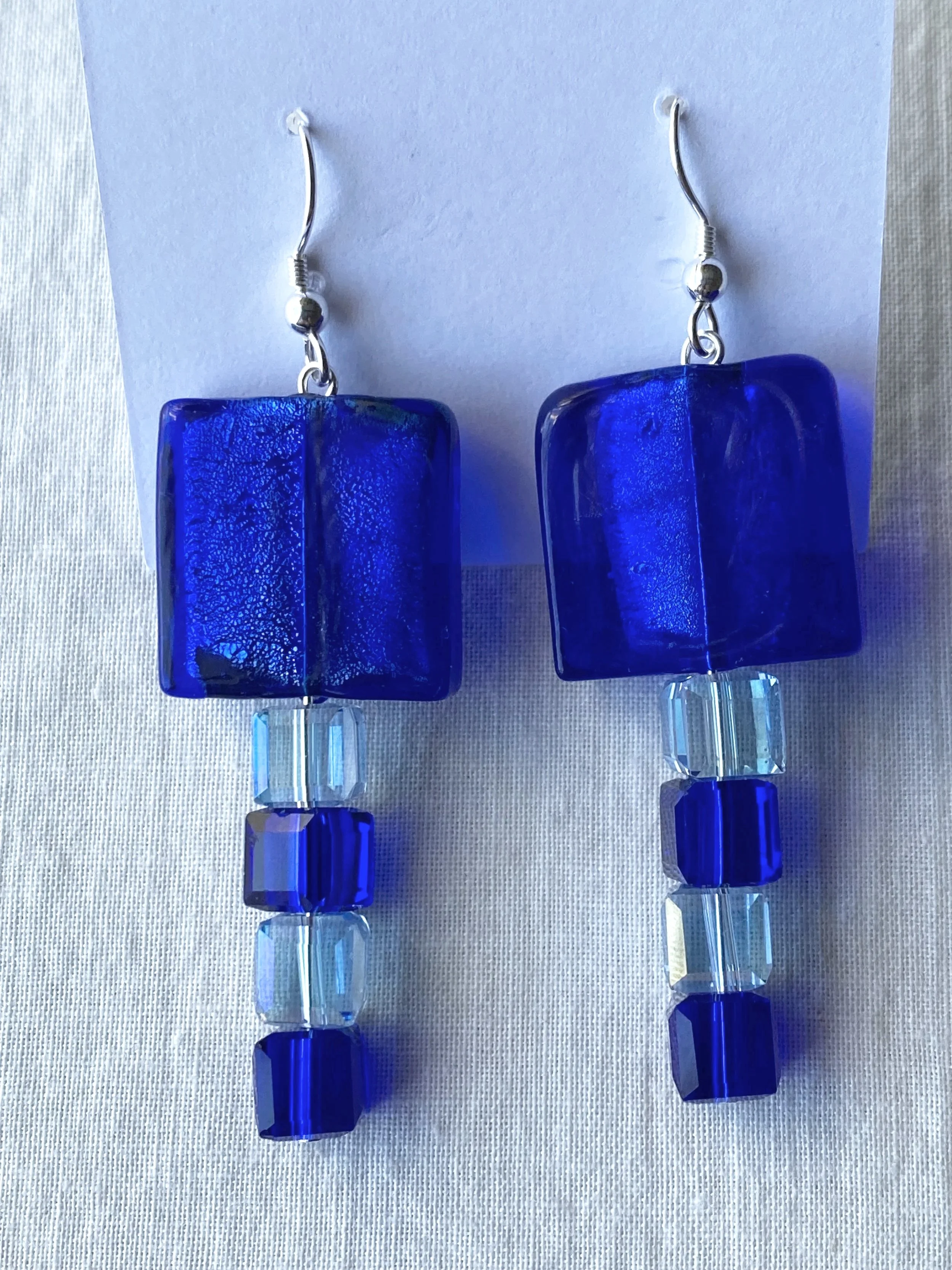 A pair of handmade blue glass dangling earrings with faceted clear beads attached to silver hooks, displayed against a white cardstock background.