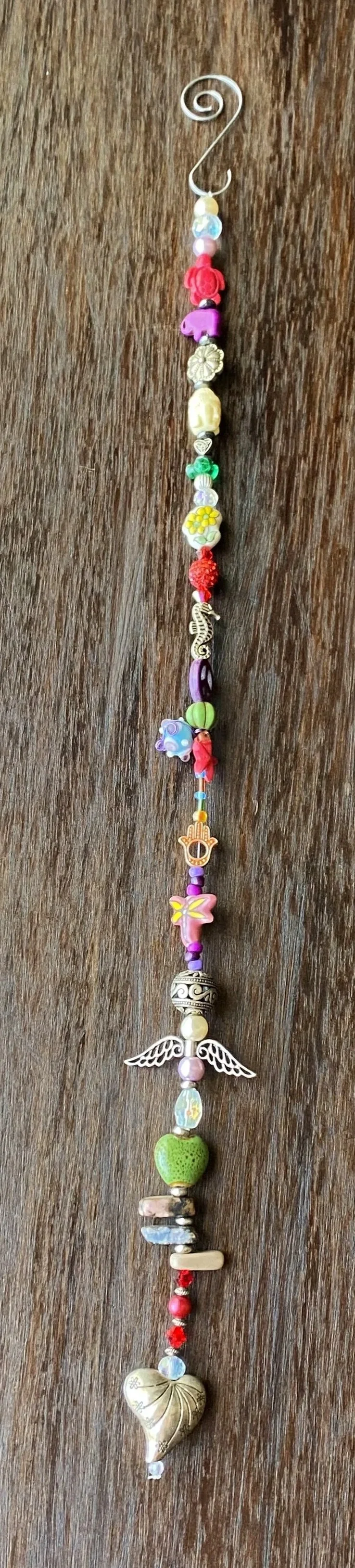Beaded jewelry piece with various colorful charms, including a seahorse, wings, hearts, flowers, animals, and abstract shapes, hanging vertically on a metal hook.