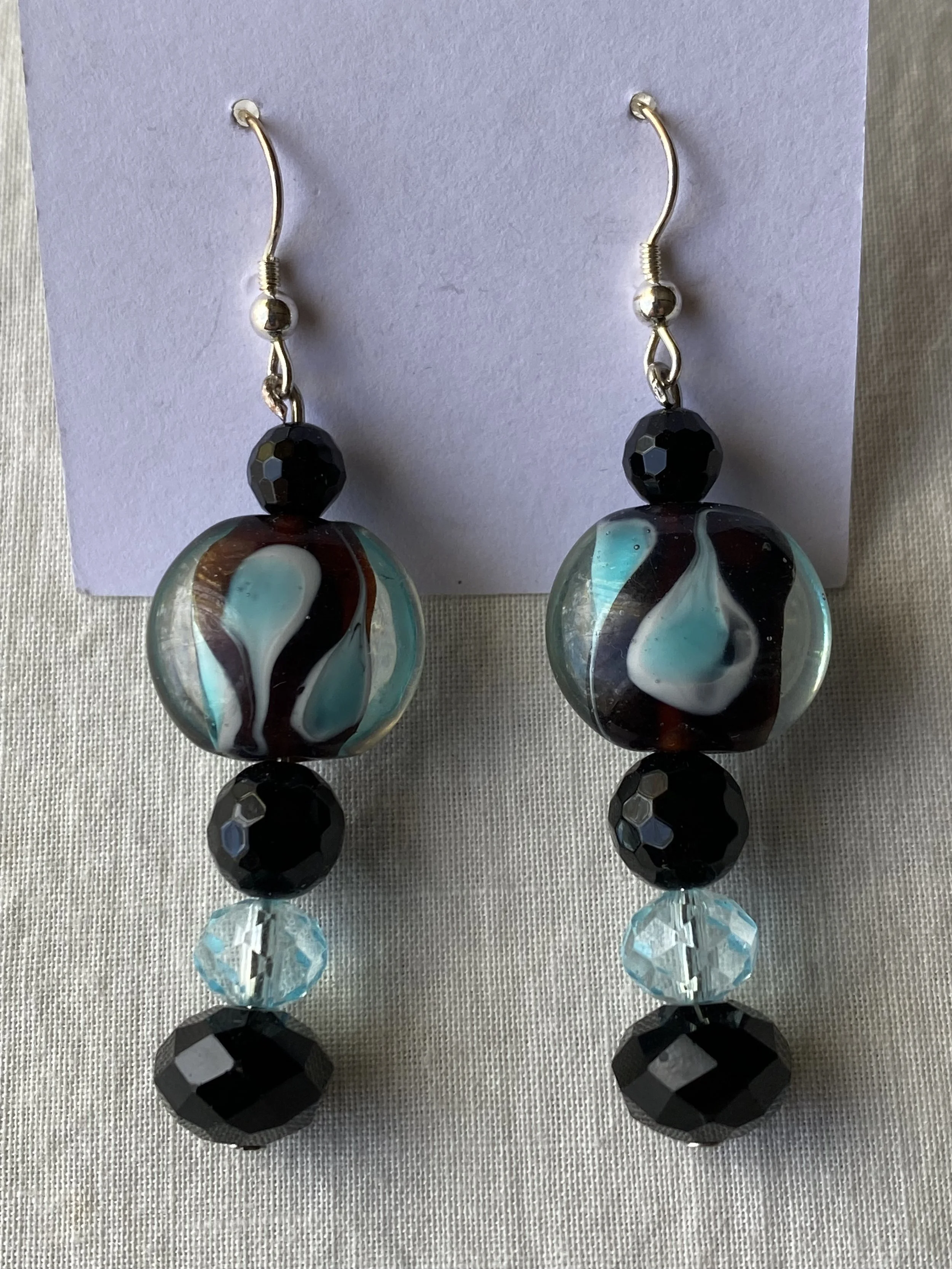 Pair of dangling earrings with large, abstract, glass beads in black, white, teal, and reddish-brown, and smaller black faceted beads above and below the glass beads, displayed on a white card.