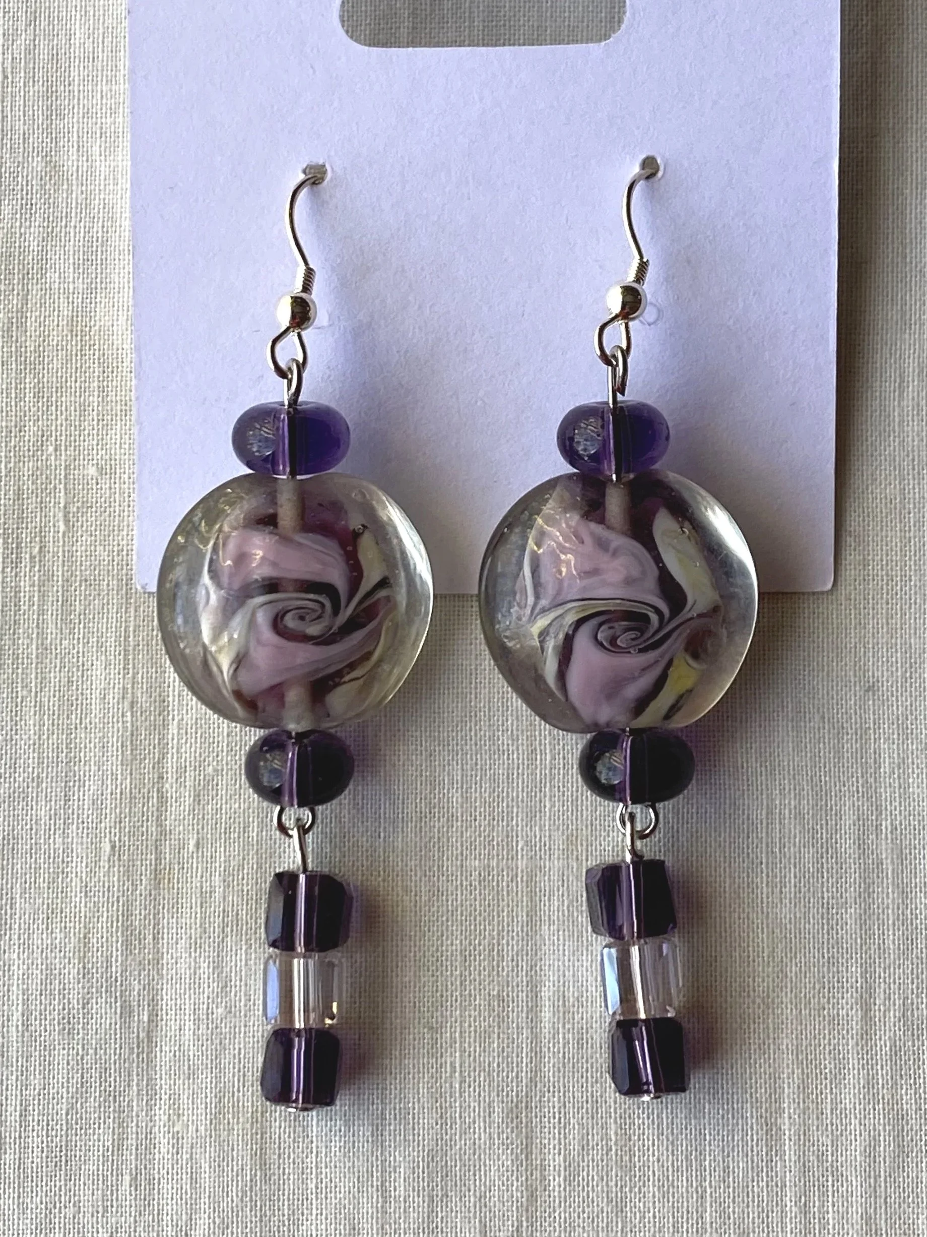 A pair of earrings with purple and black beads and swirling glass pendants, displayed on a white card against a light-colored textured background.