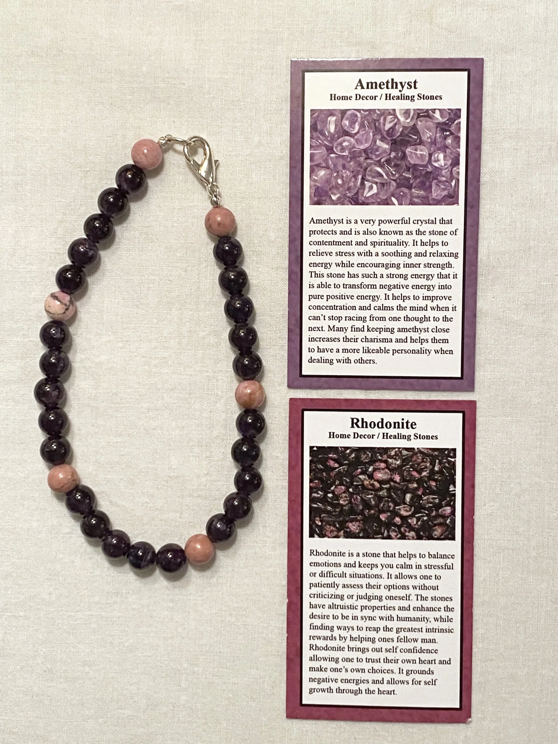 A beaded bracelet with alternating pink and dark purple beads displayed on a beige surface, next to informational cards about amethyst and rhodonite healing stones.