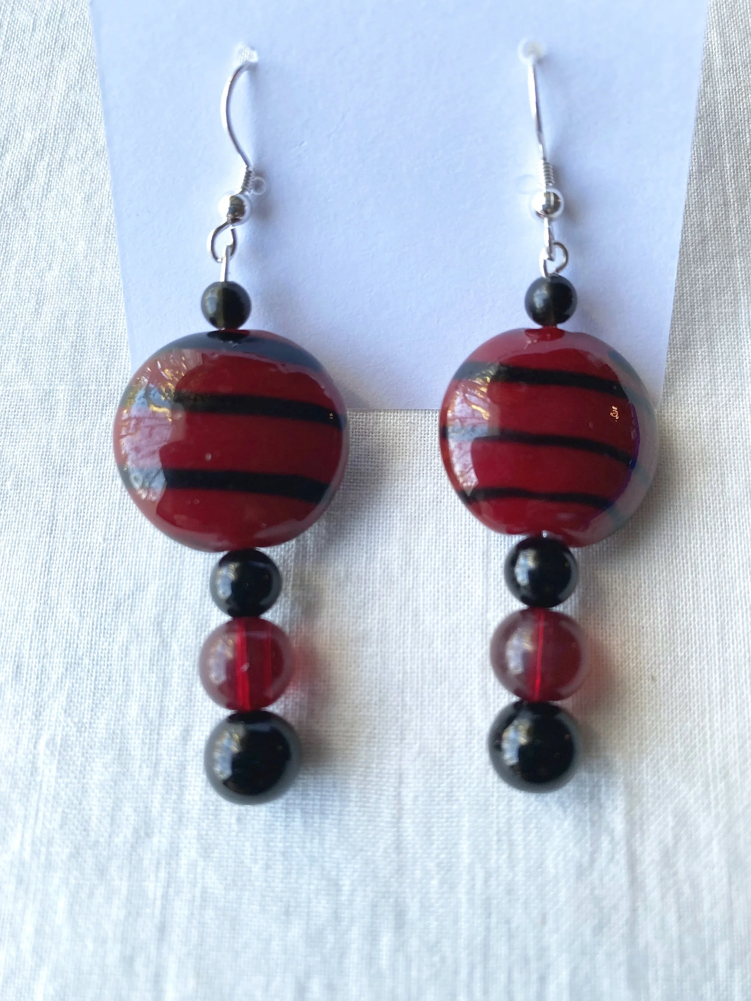 A pair of hanging earrings with large red and black striped beads, smaller black beads, and silver hooks. The earrings are displayed on a white card against a light-colored background.