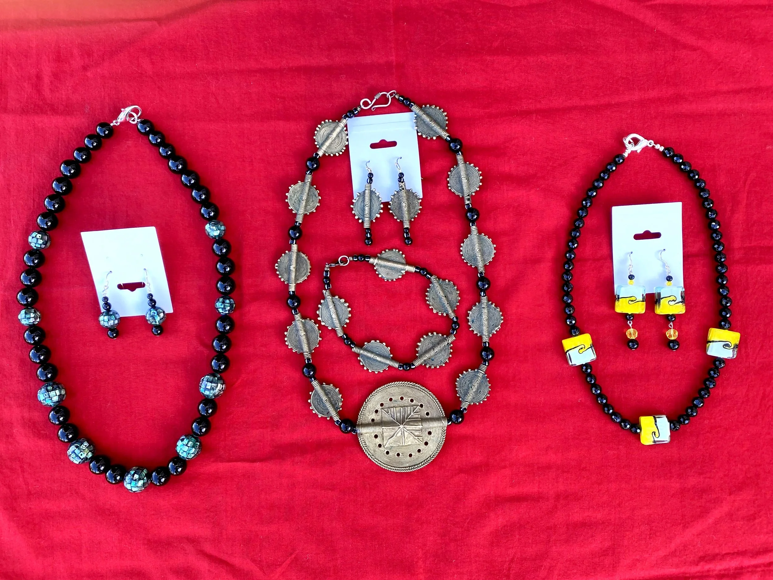 Jewelry displayed on a red fabric background, including a black beaded necklace, a silver jewelry set with circular designs, and earrings with yellow and black beads.