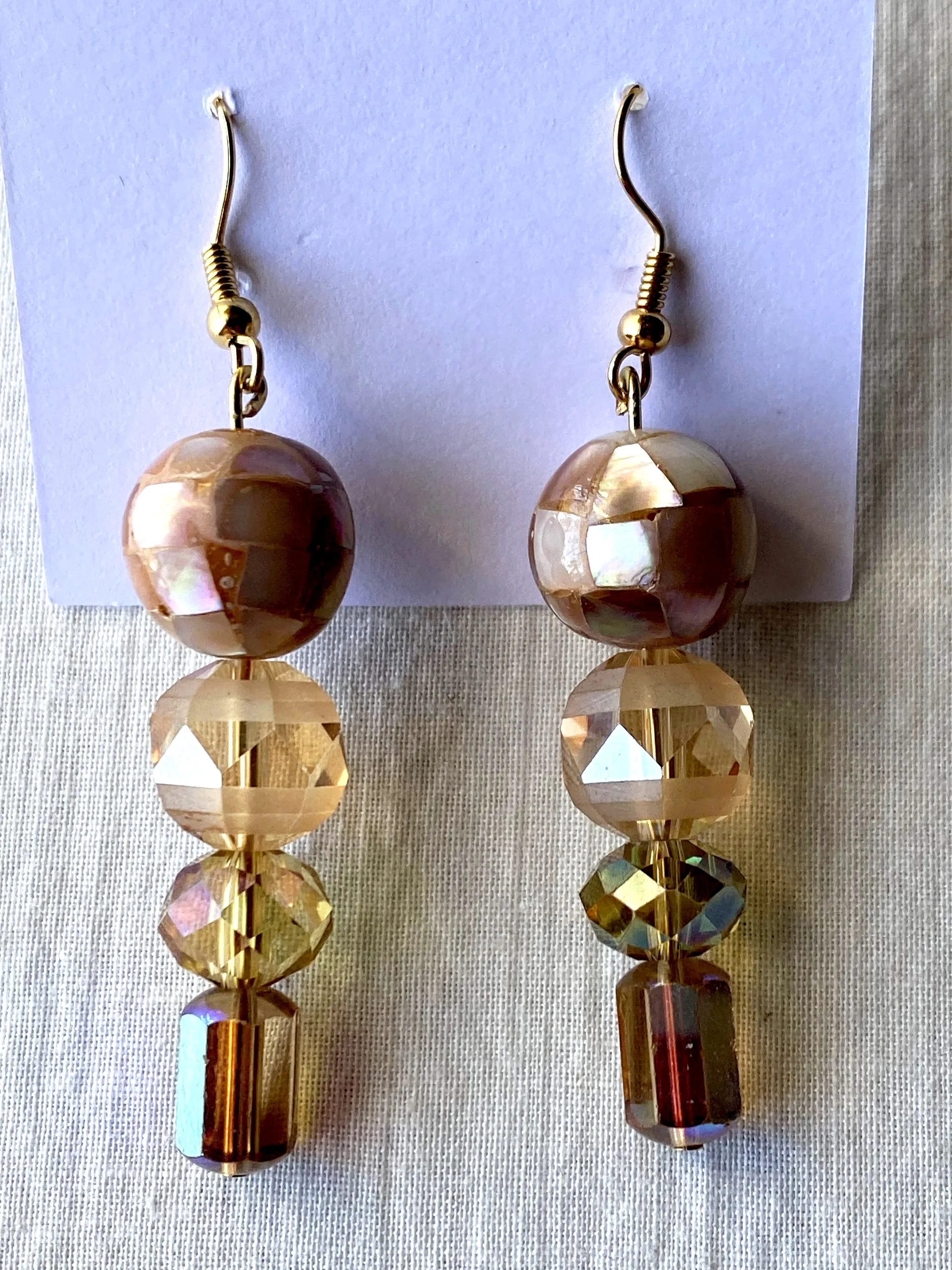 A pair of dangling earrings with round, faceted golden beads and rectangular, iridescent beads, displayed on a white card.