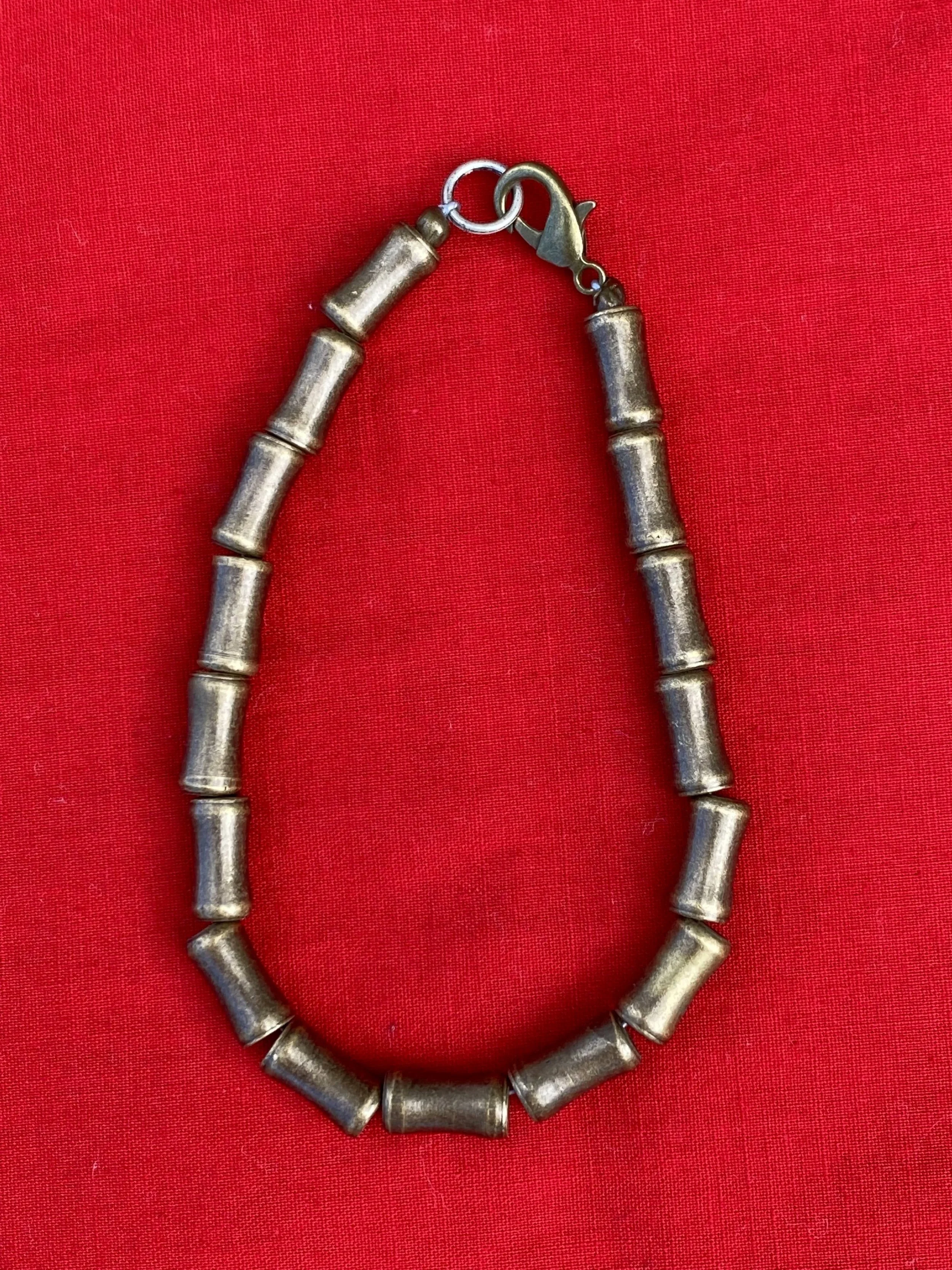 A brass bracelet made of large metallic tube-shaped beads with a lobster clasp, set against a red fabric background.