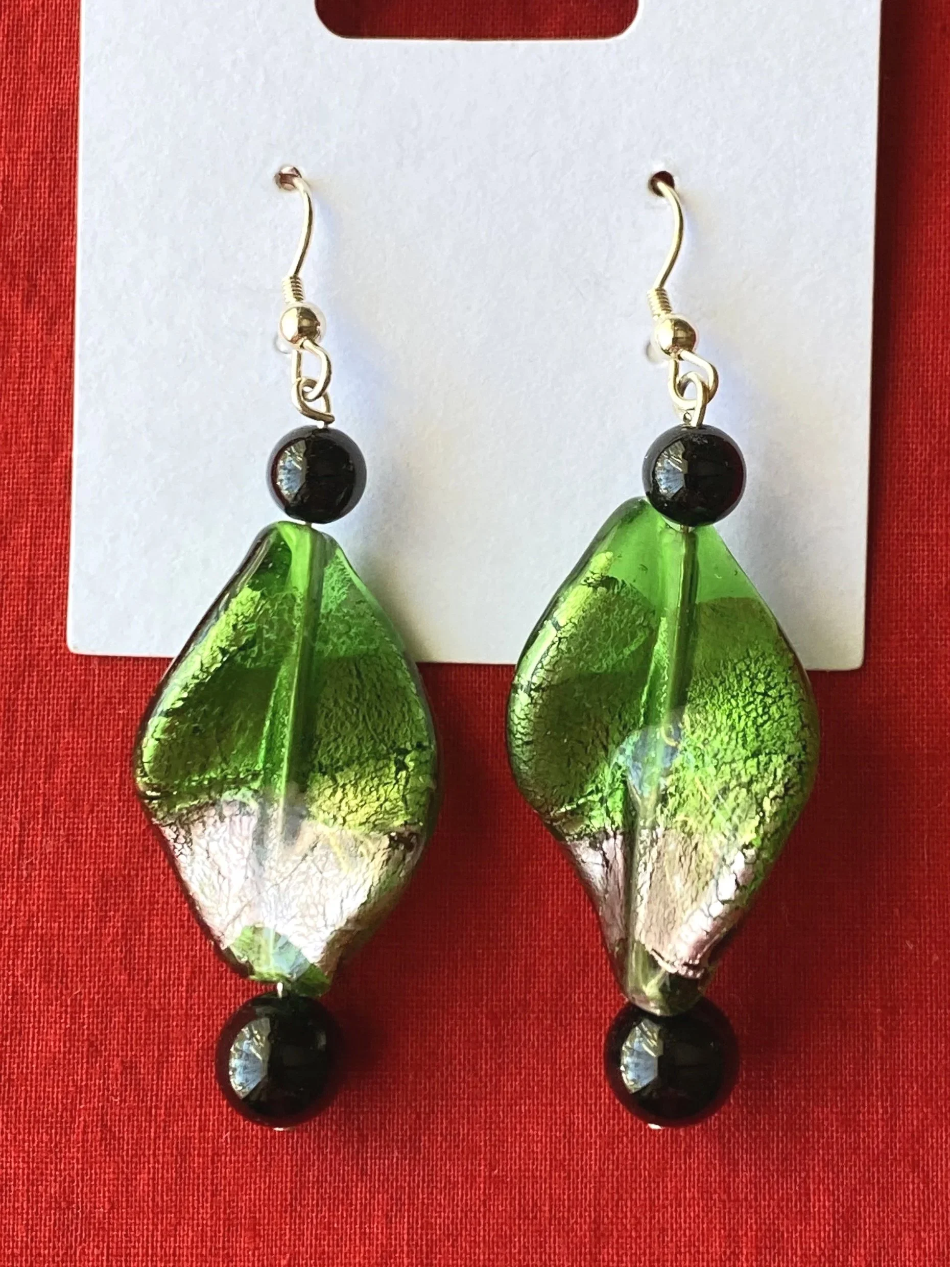 Pair of earrings with green glass leaf-shaped pendants and black beads, displayed on a red fabric background.
