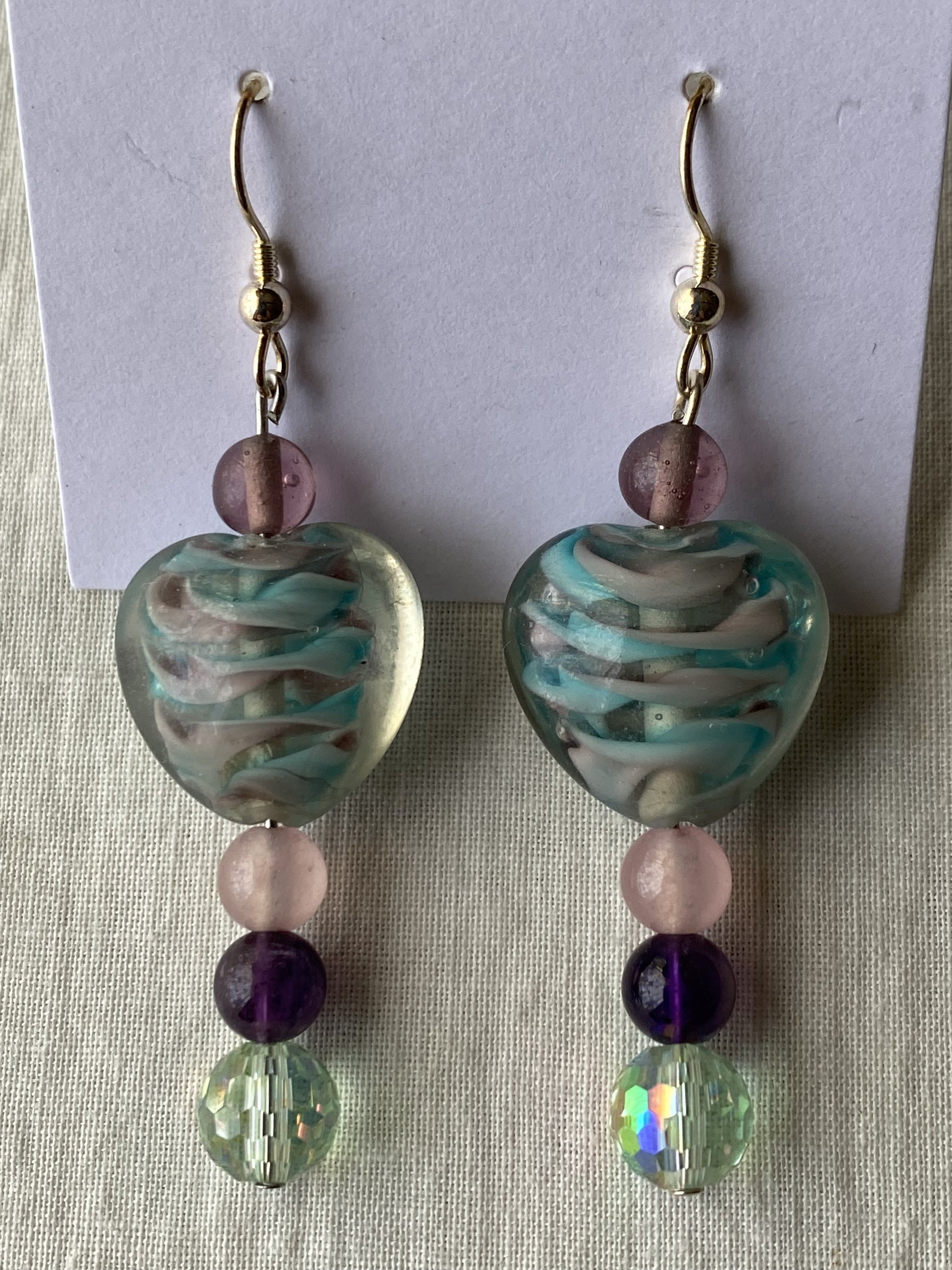 Colorful beaded earrings with round and faceted beads in purple, pink, and iridescent colors, displayed on a white card.
