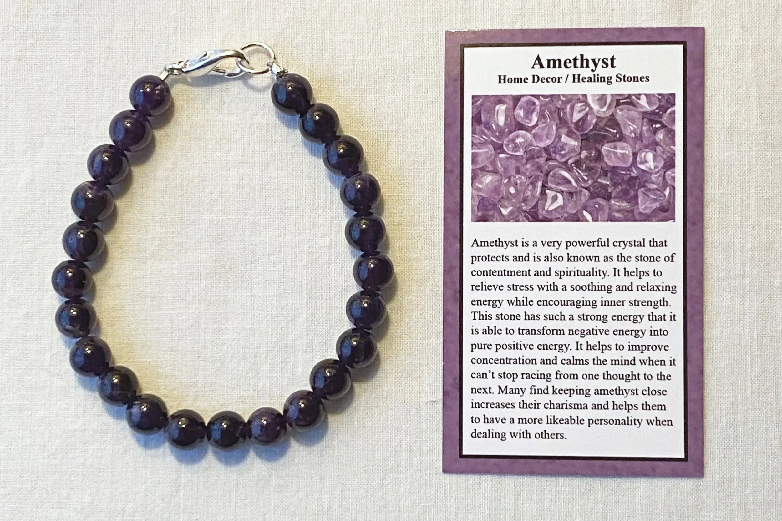 Close-up of a bracelet with dark purple amethyst beads with a silver clasp, lying on a cream-colored surface next to an informational card about amethyst healing stones.