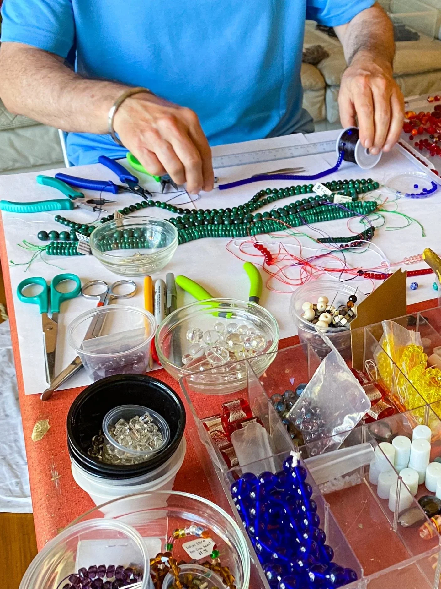 Jeff is in a blue shirt is working at a table with various beads, jewelry-making tools, and supplies. He is creating jewelry with beads.