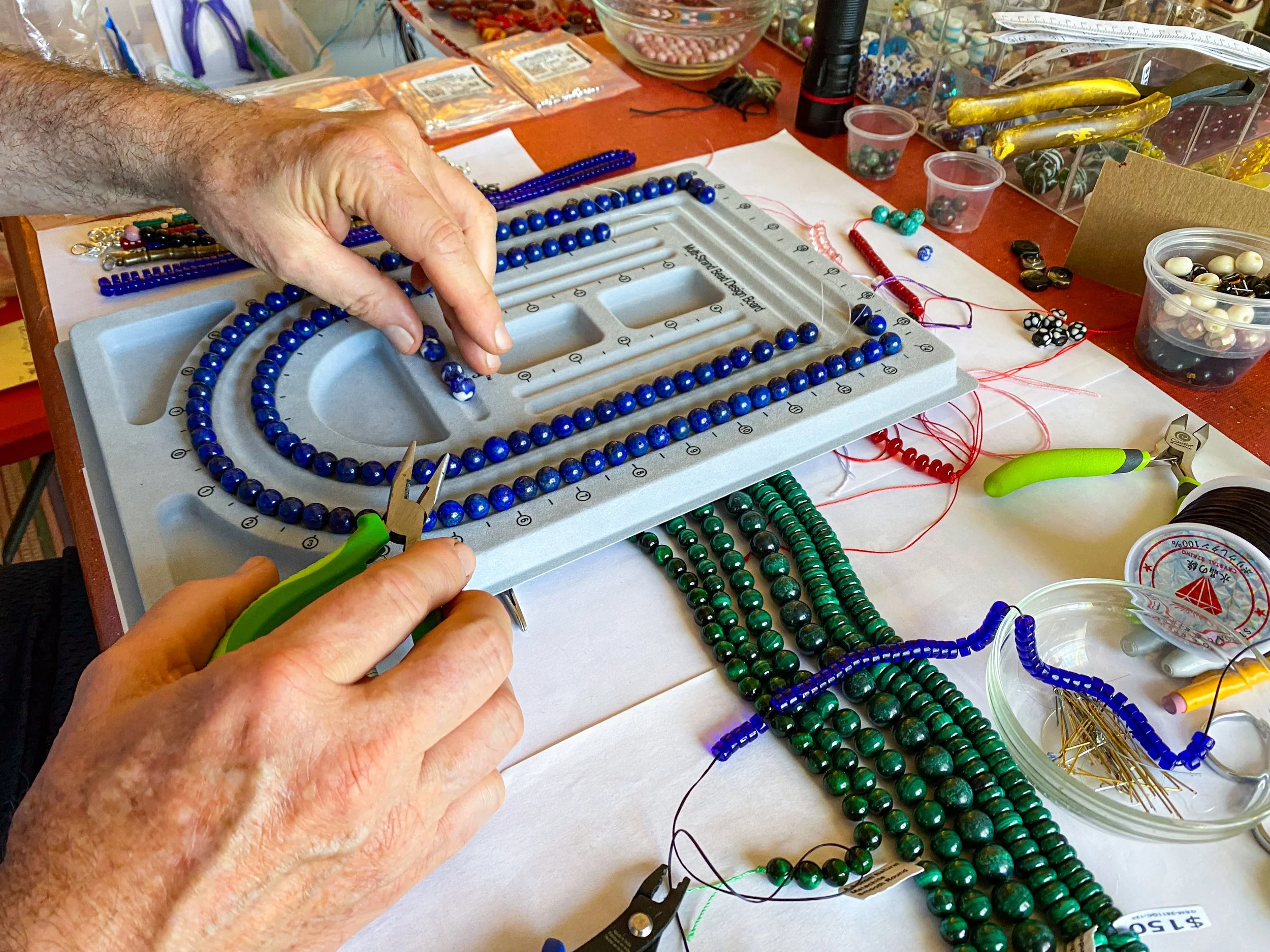 Close-up of a Jeff stringing blue and green beads on a beading board, surrounded by various beads, tools, and craft supplies on a work table.