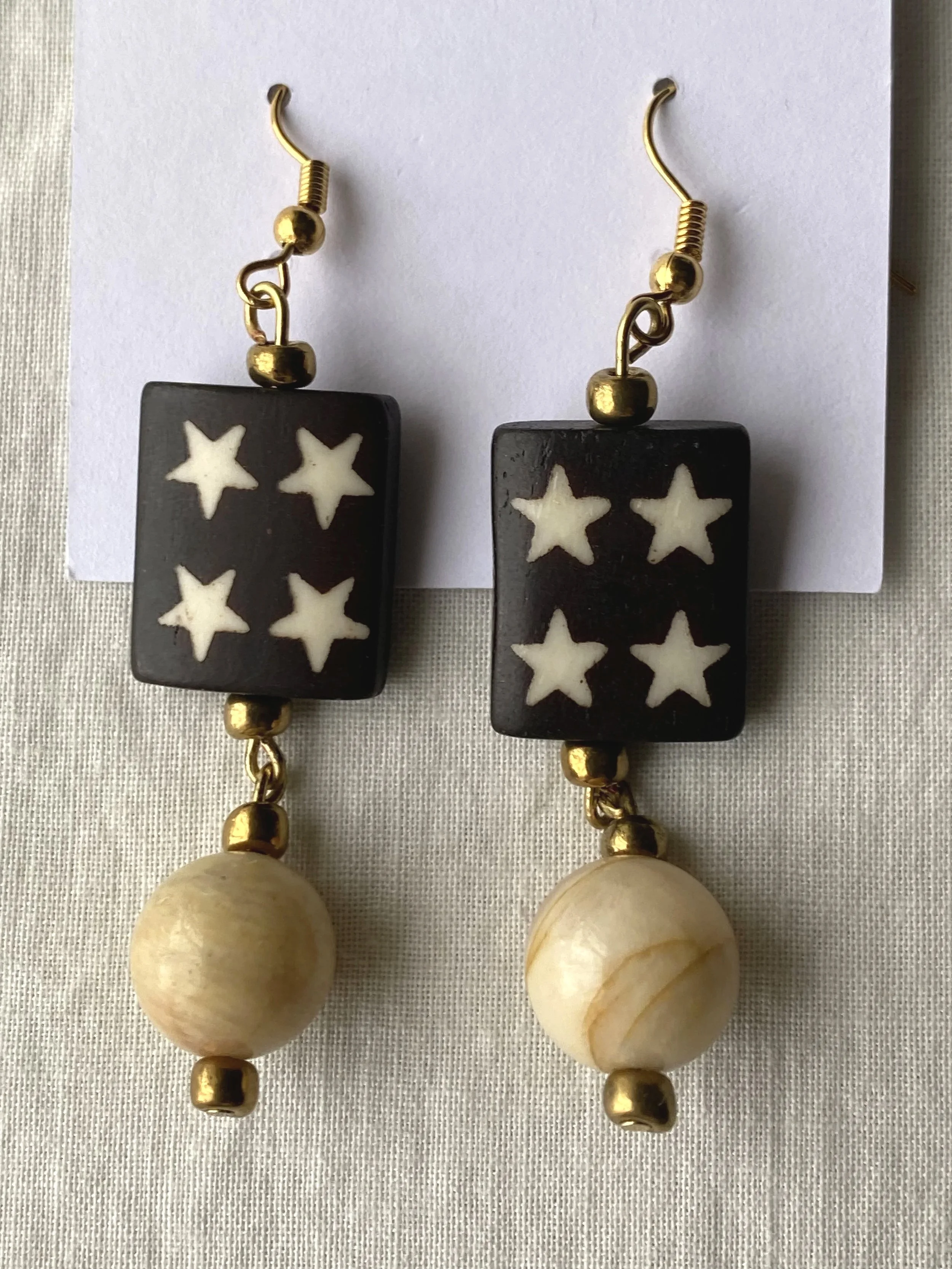 A pair of earrings featuring black rectangular beads with white star patterns, gold beads, and large round ivory-colored beads, displayed on a white background.