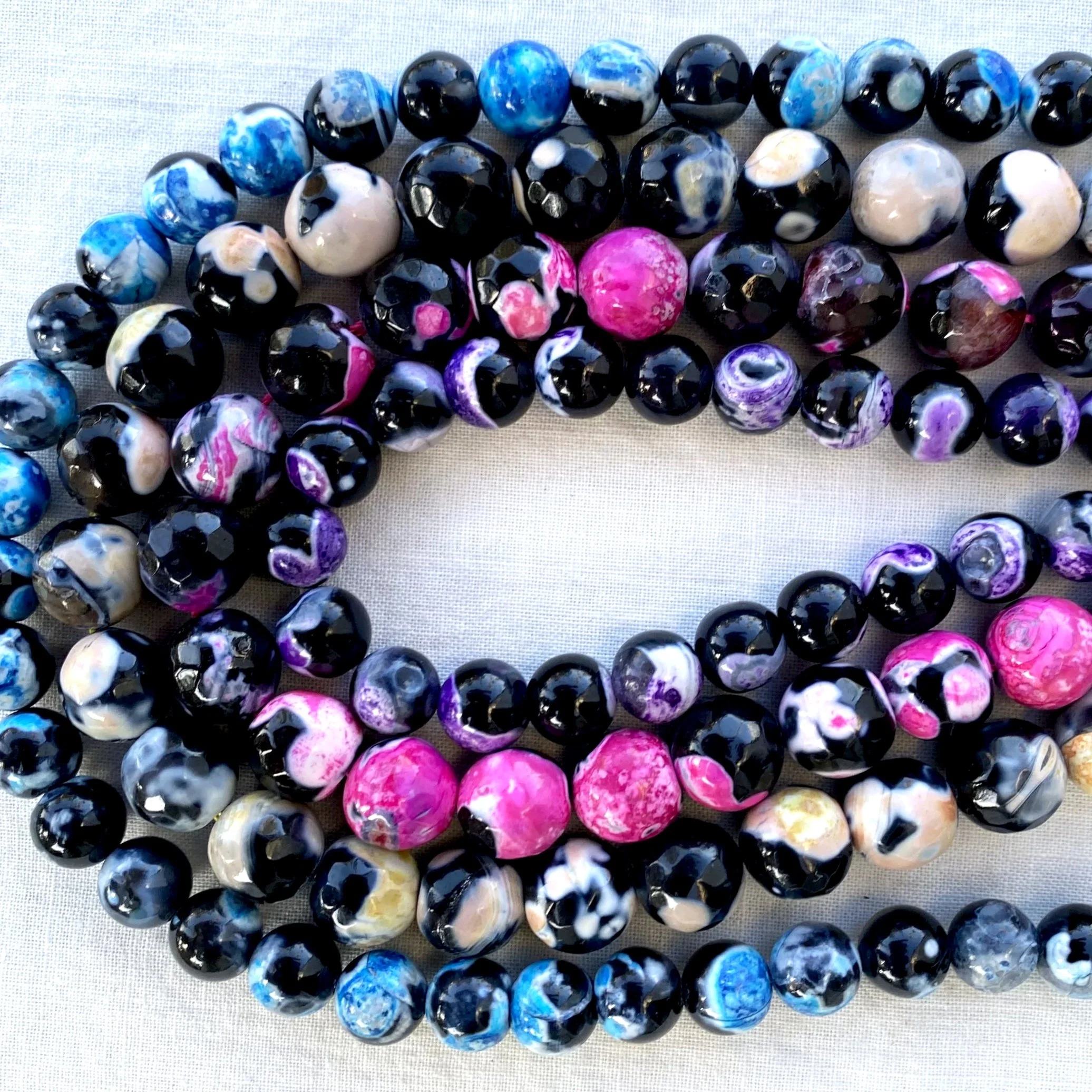 Close-up of several strands of marbled glass beads in shades of blue, black, white, purple, and pink, arranged on a light-colored fabric background.