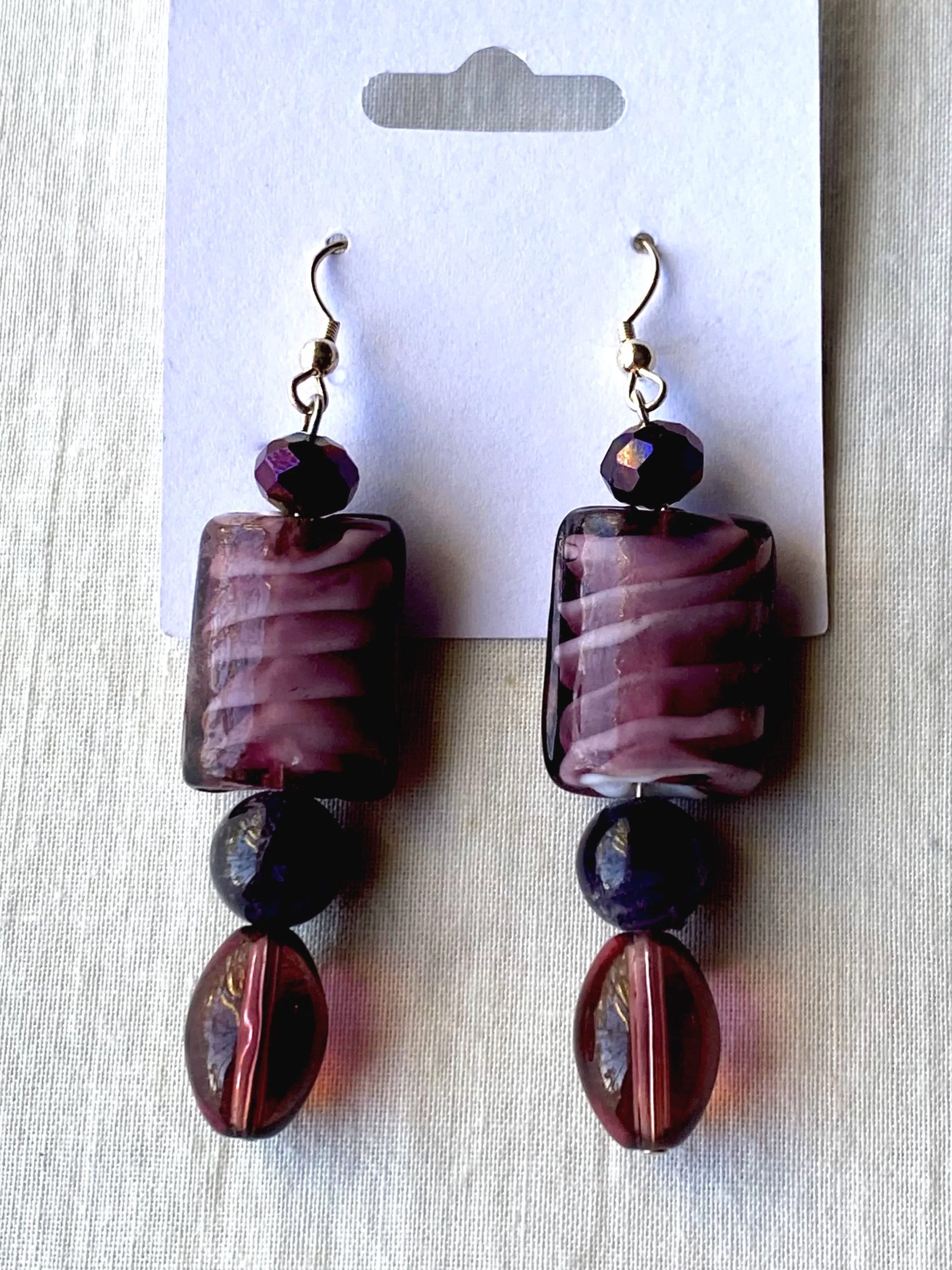 Pair of earrings featuring purple and black beads with large rectangular purple marbled pendants, displayed on white card.