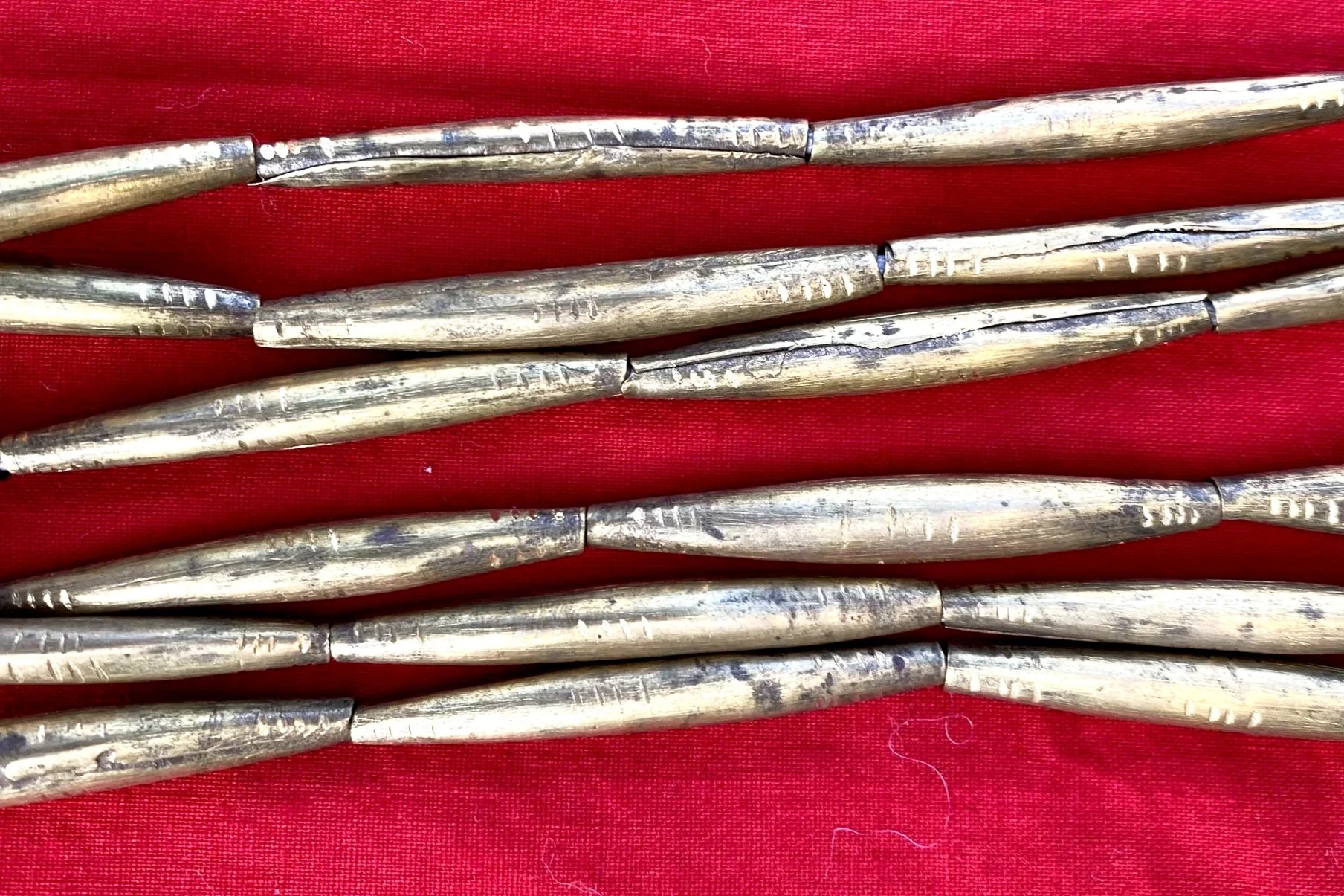 Multiple elongated, brass metallic beads with worn, rough surfaces, laid out on a red fabric background.