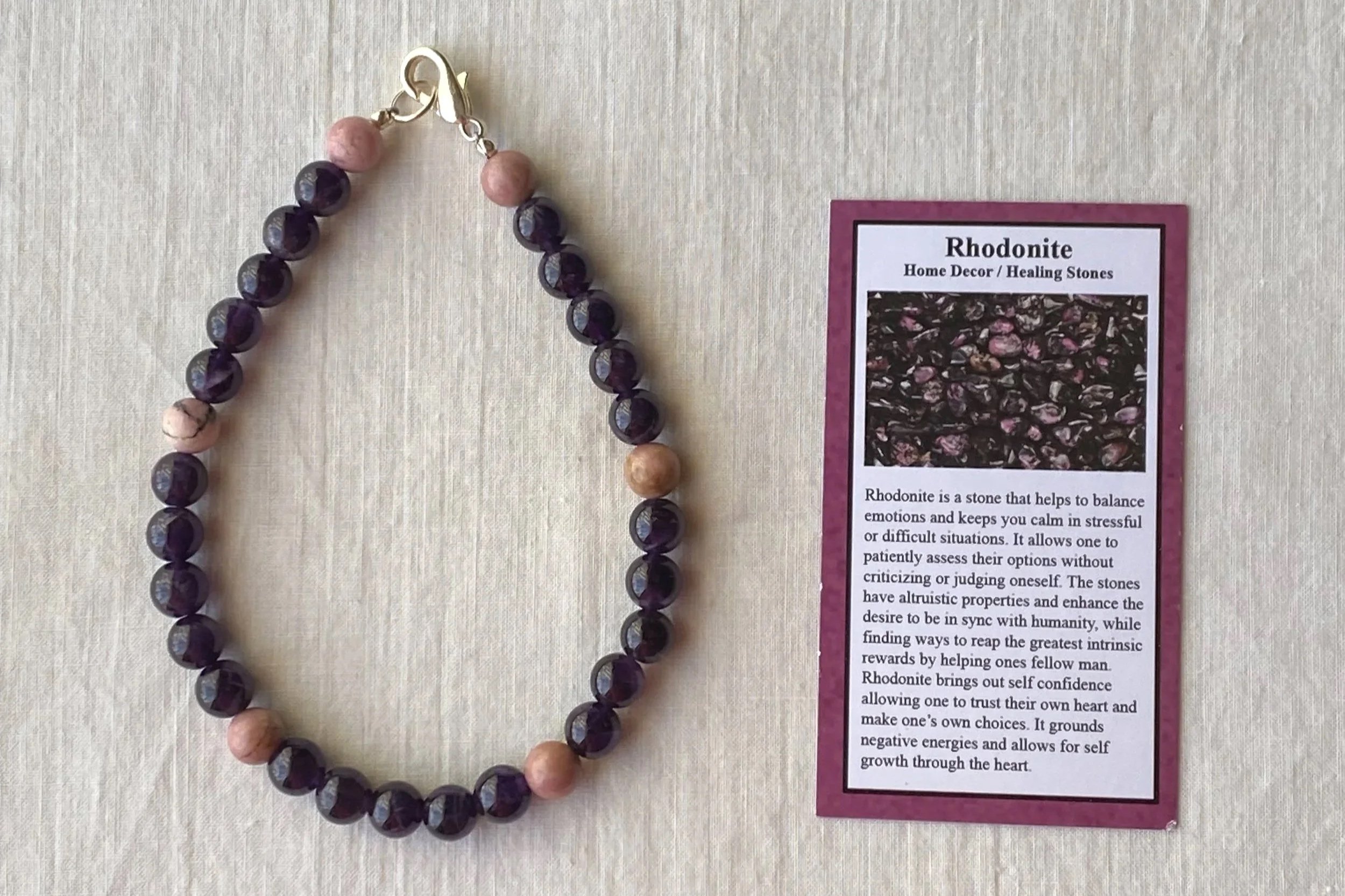 A beaded bracelet with purple and pink stones displayed on a light-colored surface next to a card describing Rhodonite as a healing stone. The card mentions Rhodonite's properties related to balancing emotions and promoting self-confidence.