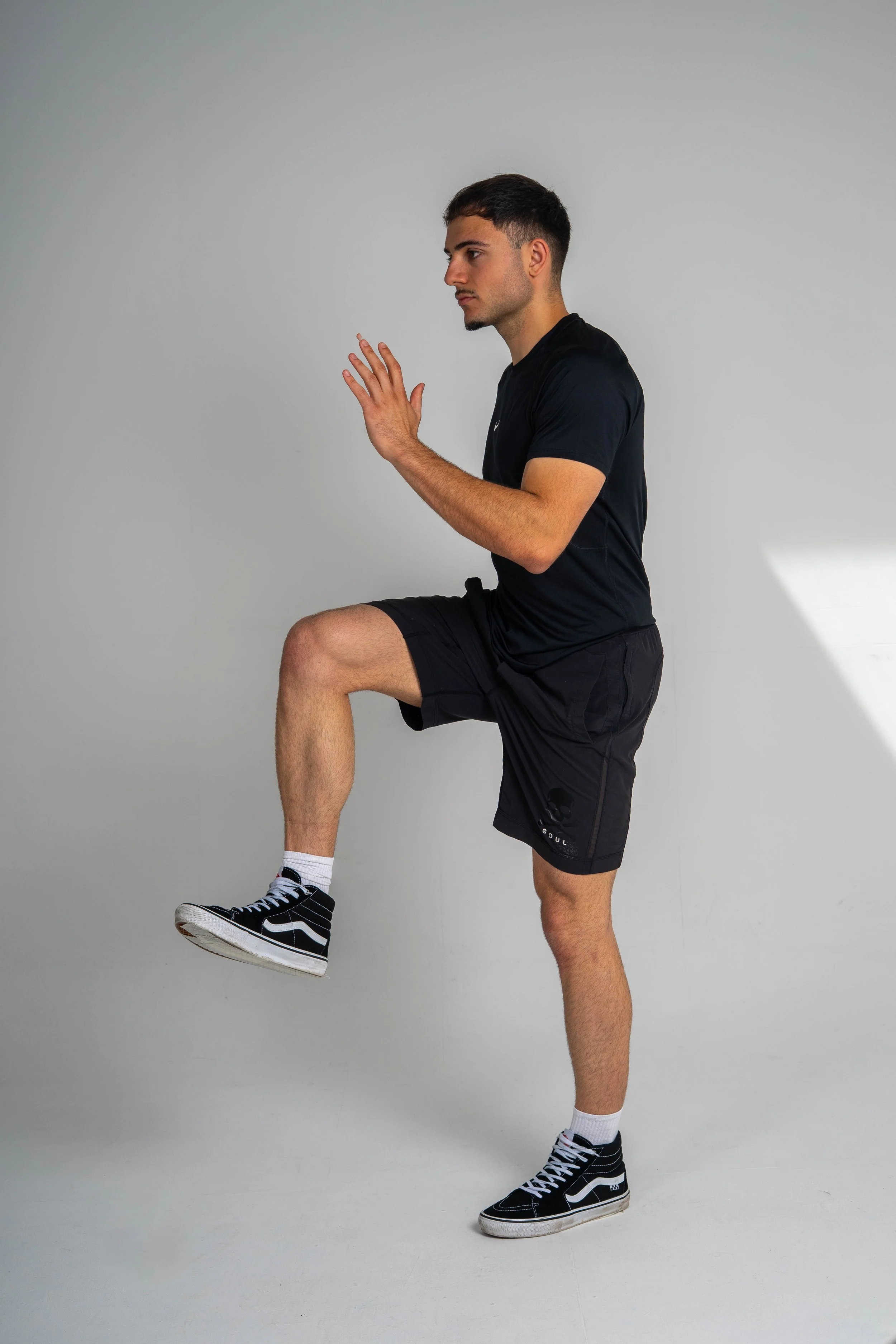 A young man performing a balance exercise, standing on one leg with the other knee raised, and holding his hands together in front of his chest, against a plain gray background.