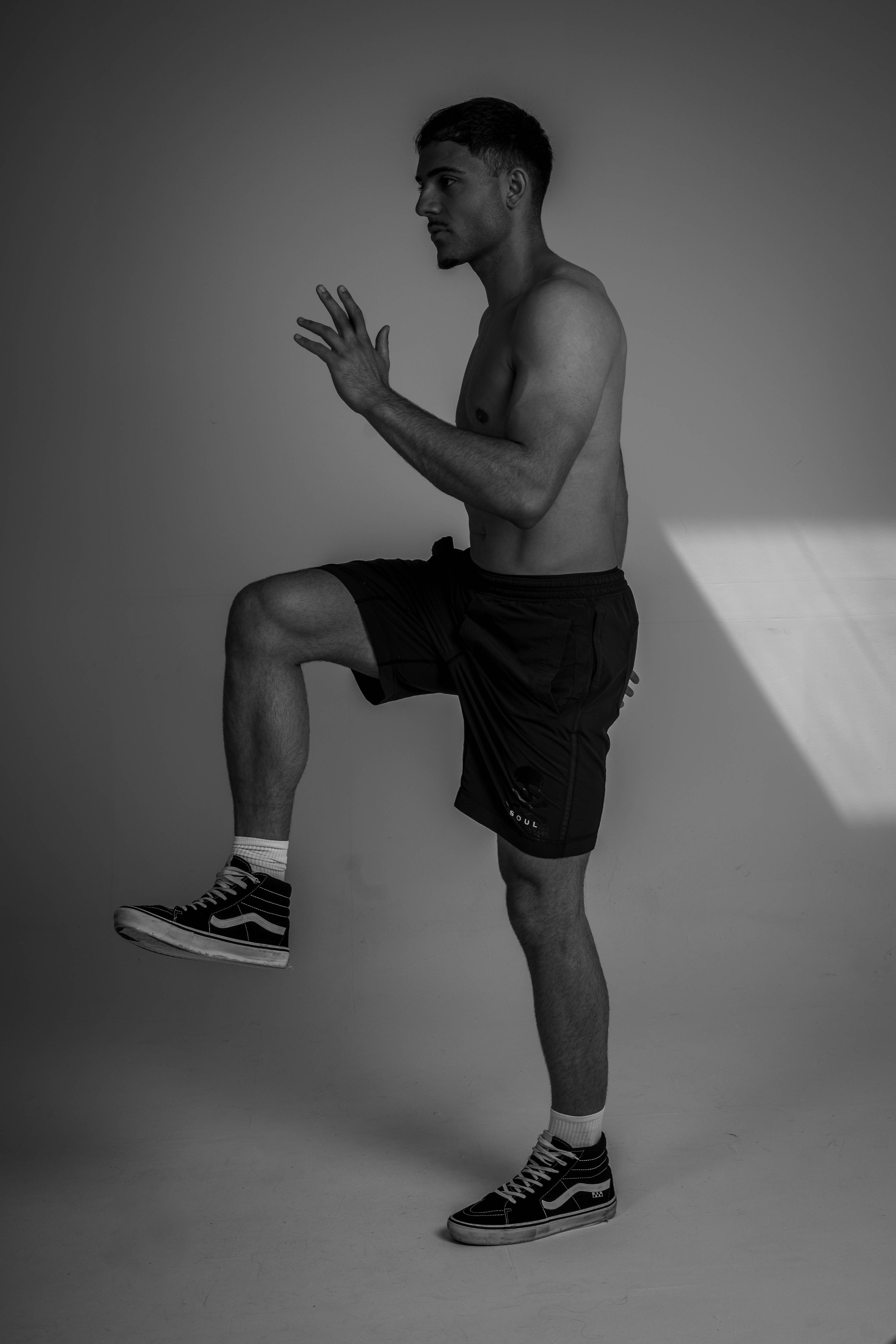A young man with short hair, shirtless, wearing black shorts, high-top sneakers, and socks, performing a high-knee exercise indoors against a plain background.