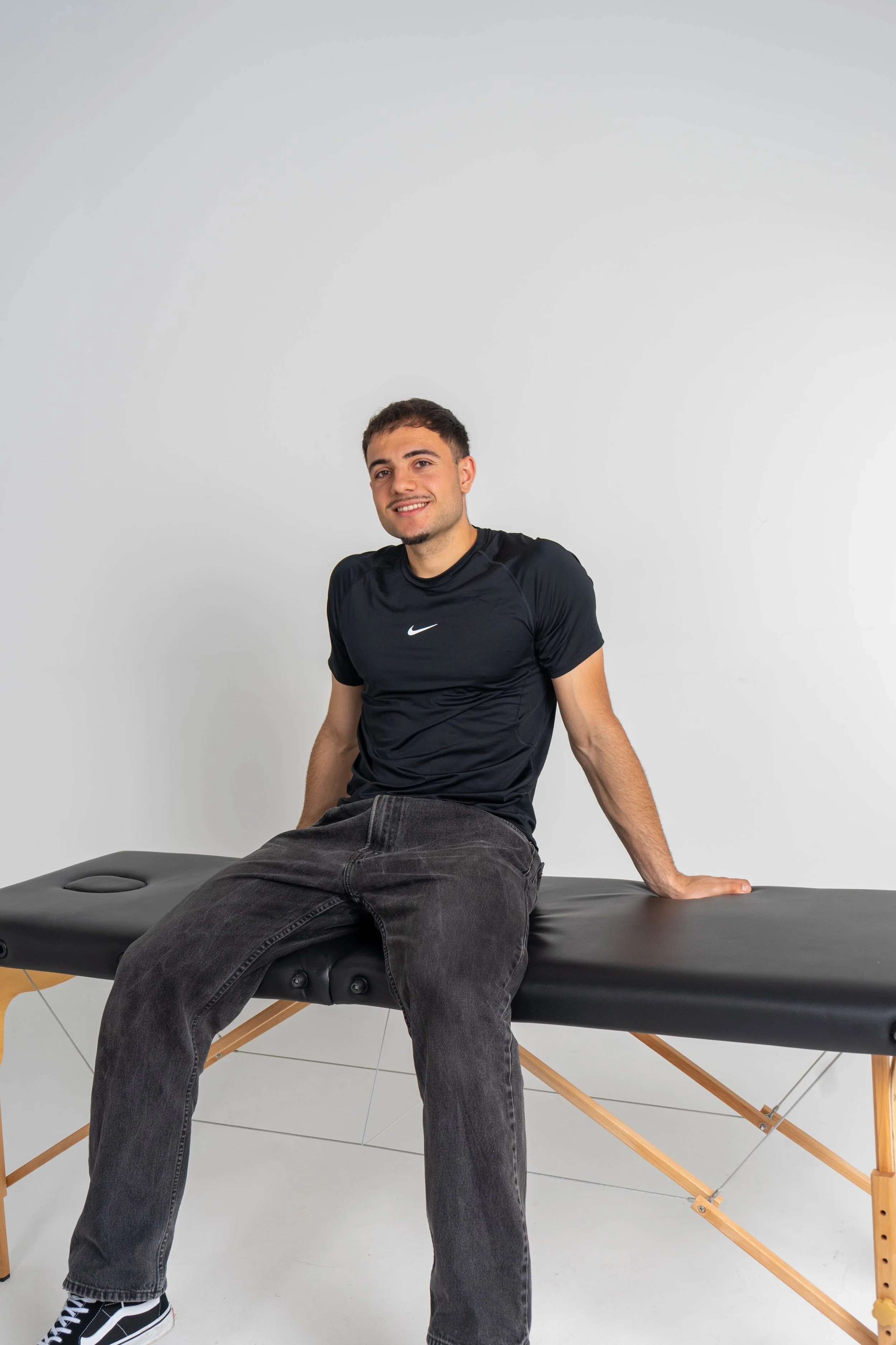 A young man with short dark hair and a light beard sitting on a black massage or therapy table with a wooden frame, smiling at the camera, against a plain light gray wall.