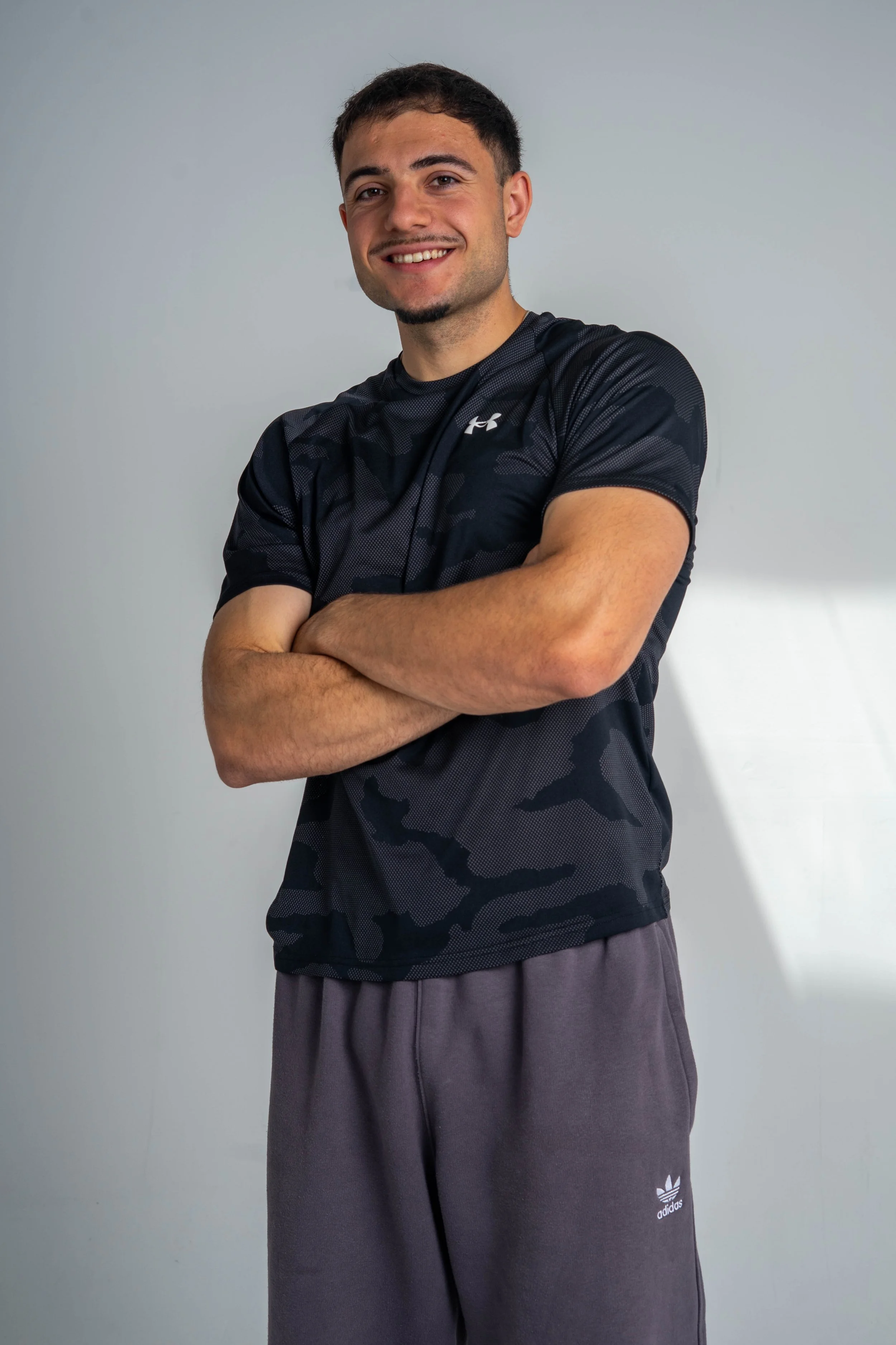 A smiling young man with crossed arms, wearing a black Under Armour athletic shirt and gray Adidas sweatpants in front of a plain light gray background.