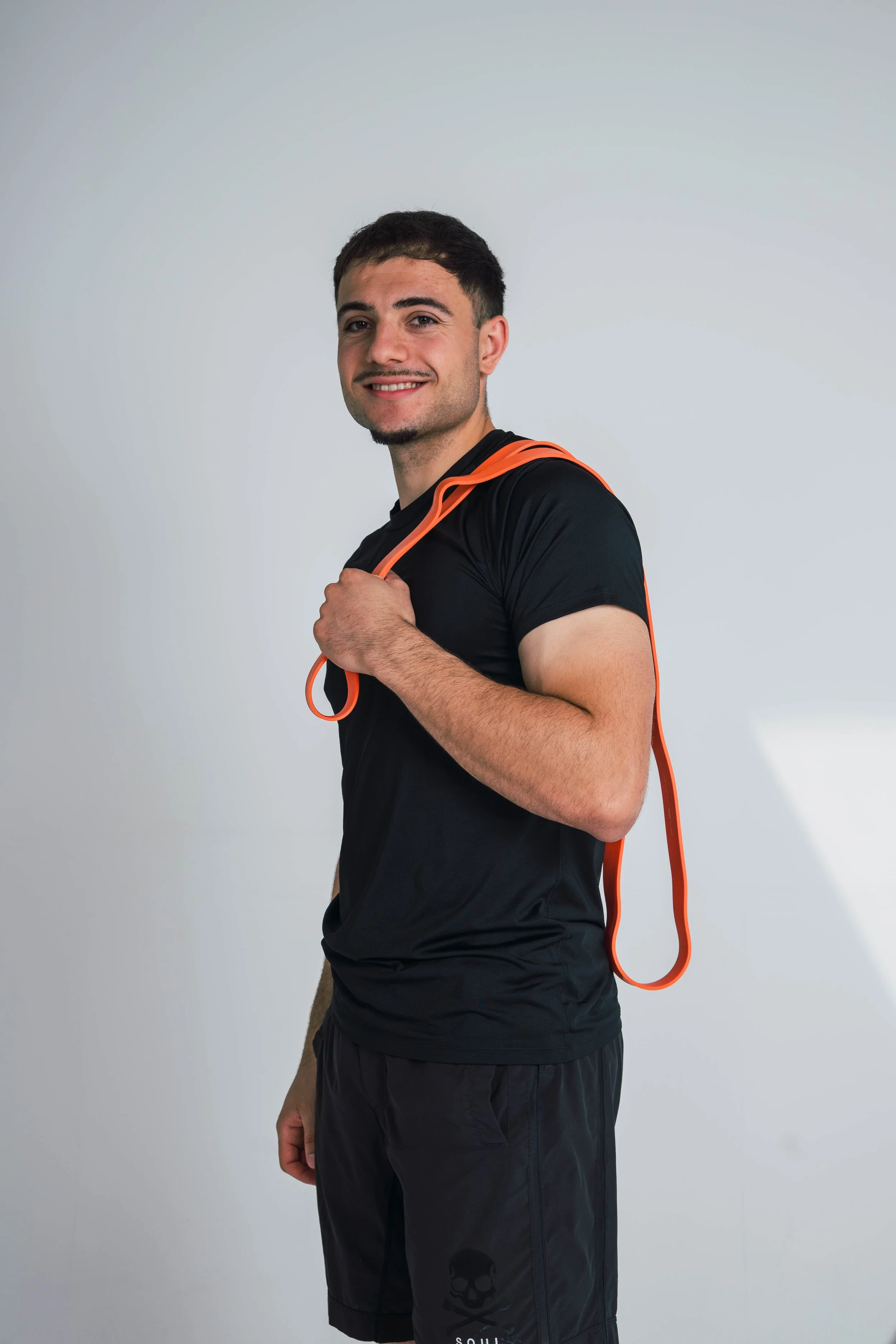 A young man with short dark hair and light skin smiling at the camera, wearing a black athletic shirt and black shorts with a skull logo, holding an orange jump rope over his shoulder against a plain light grey background.
