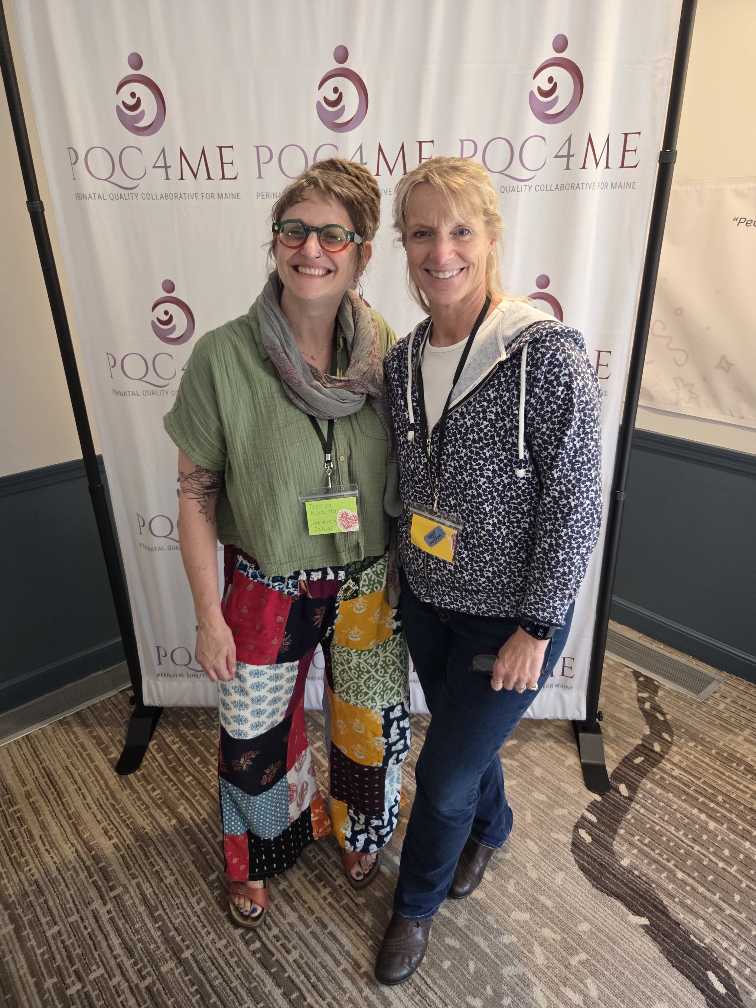 Two women standing together, smiling, in front of a promotional backdrop for PQC4ME, a collaborative organization for Maine. One woman has short, curly hair, glasses, and tattoos on her arm, wearing a green top and colorful patchwork pants. The other has shoulder-length blonde hair, wearing a navy patterned jacket and jeans.