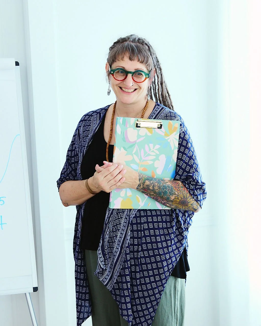 A smiling woman with glasses, dreadlocks, and tattoos, holding a clipboard, standing in a bright room.