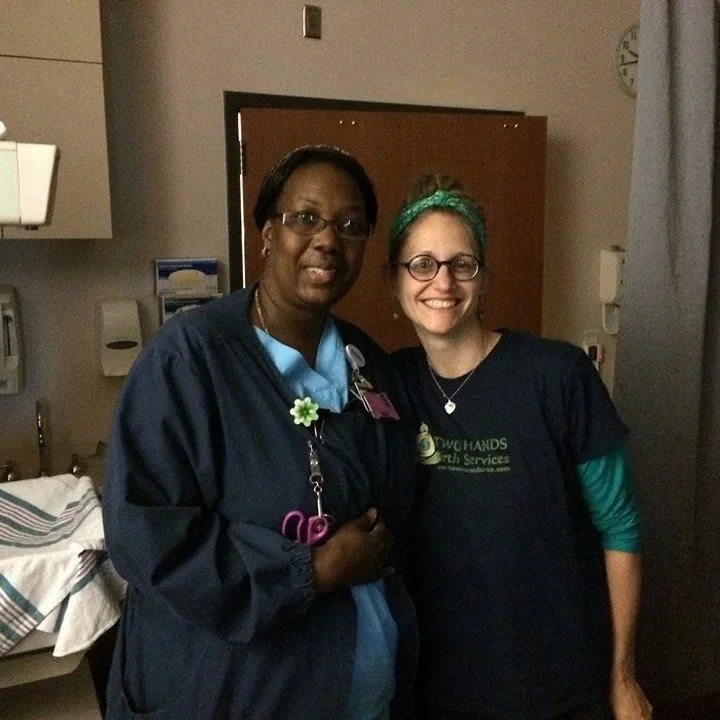 Two women standing together in a hospital room, smiling at the camera. One is a nurse in dark blue scrubs, and the other is a doula wearing a black T-shirt with a logo.