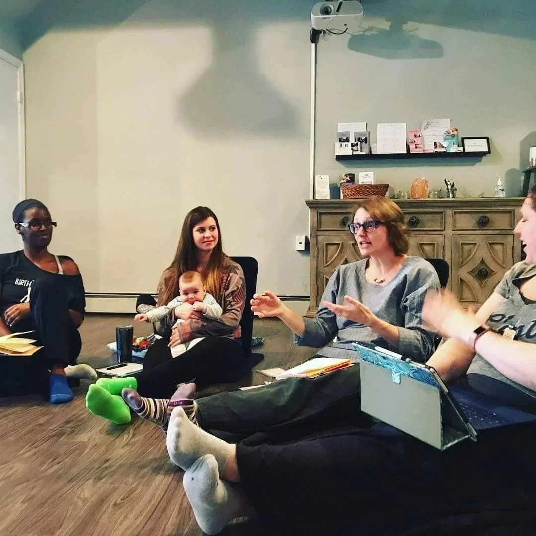 A group of women sitting on the floor in a circle, having a discussion, with one woman holding a baby, in a room with a bookshelf and decorative items.