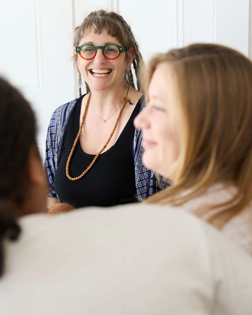 A woman with bangs and dreadlocks, colorful glasses, and earrings, smiling at a couple in a bright room.