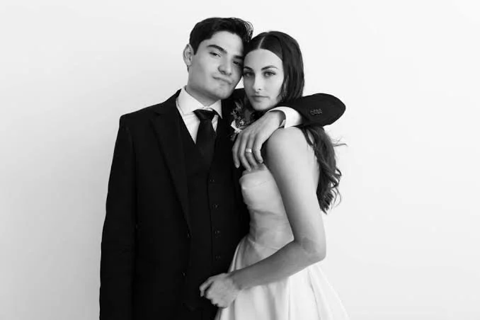 A young couple dressed in formal attire, with the man wearing a suit and tie, and the woman in a sleeveless dress, standing close together against a plain background.