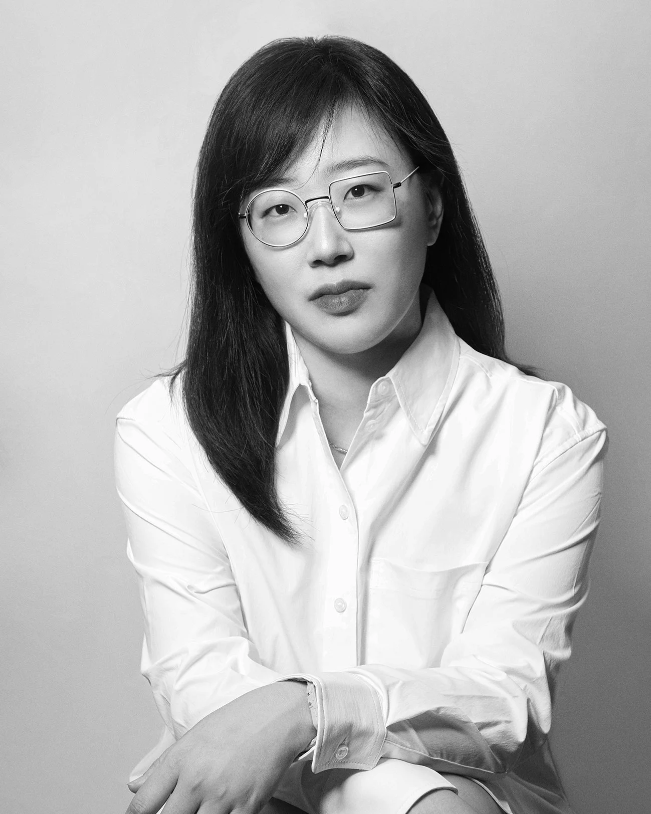Black and white portrait of an Asian woman with long dark hair and glasses, wearing a white button-up shirt, sitting with crossed arms against a plain background.