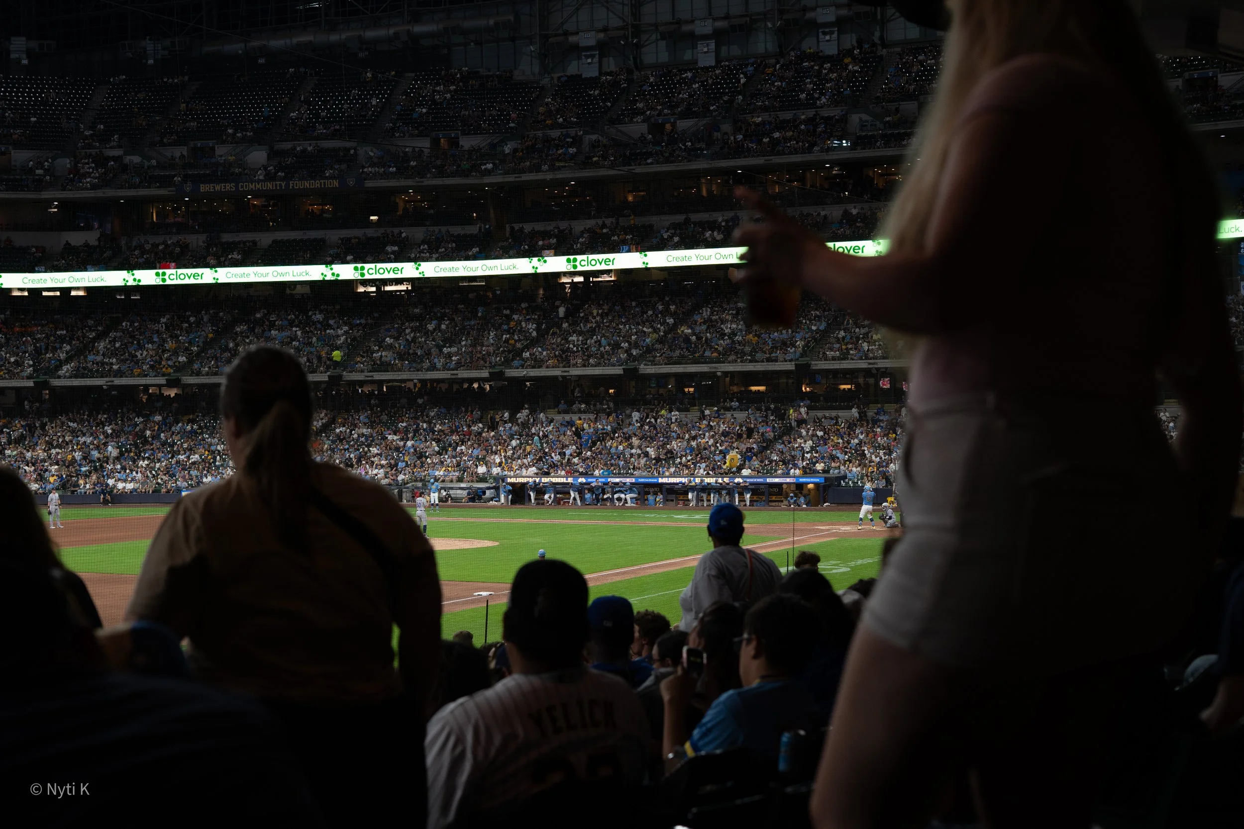 View of a baseball stadium filled with spectators, some are standing and watching the game. In the foreground, there are silhouettes of fans, including a person with a blonde ponytail, pointing towards the field. The field is visible with players positioned for a play, and a digital advertisement banner for 'Clover' with the slogan 'Create Your Own Luck' is displayed across the middle of the stadium.