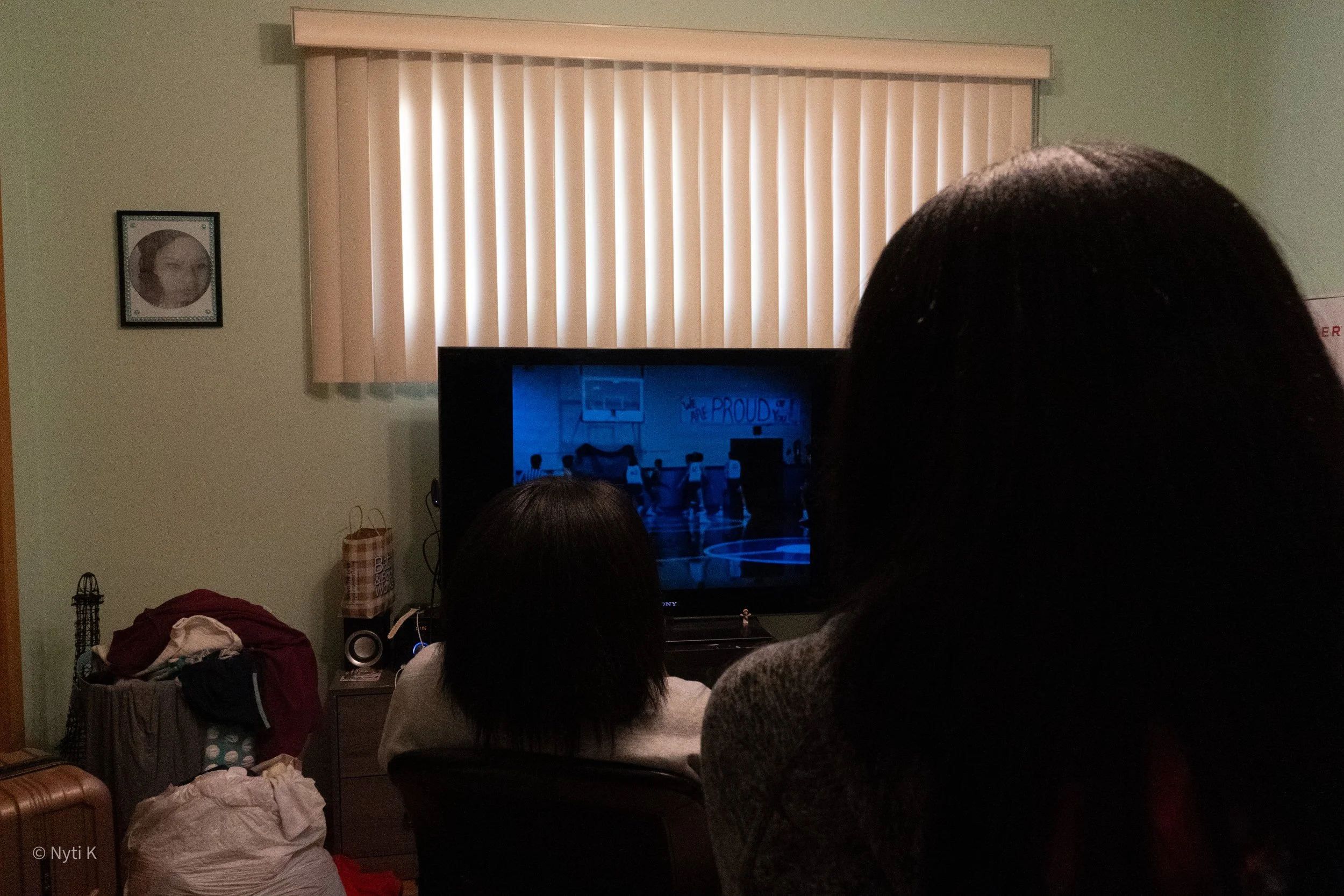 People sitting in a living room, watching a sports game on television, with vertical blinds covering the window and some household items visible.