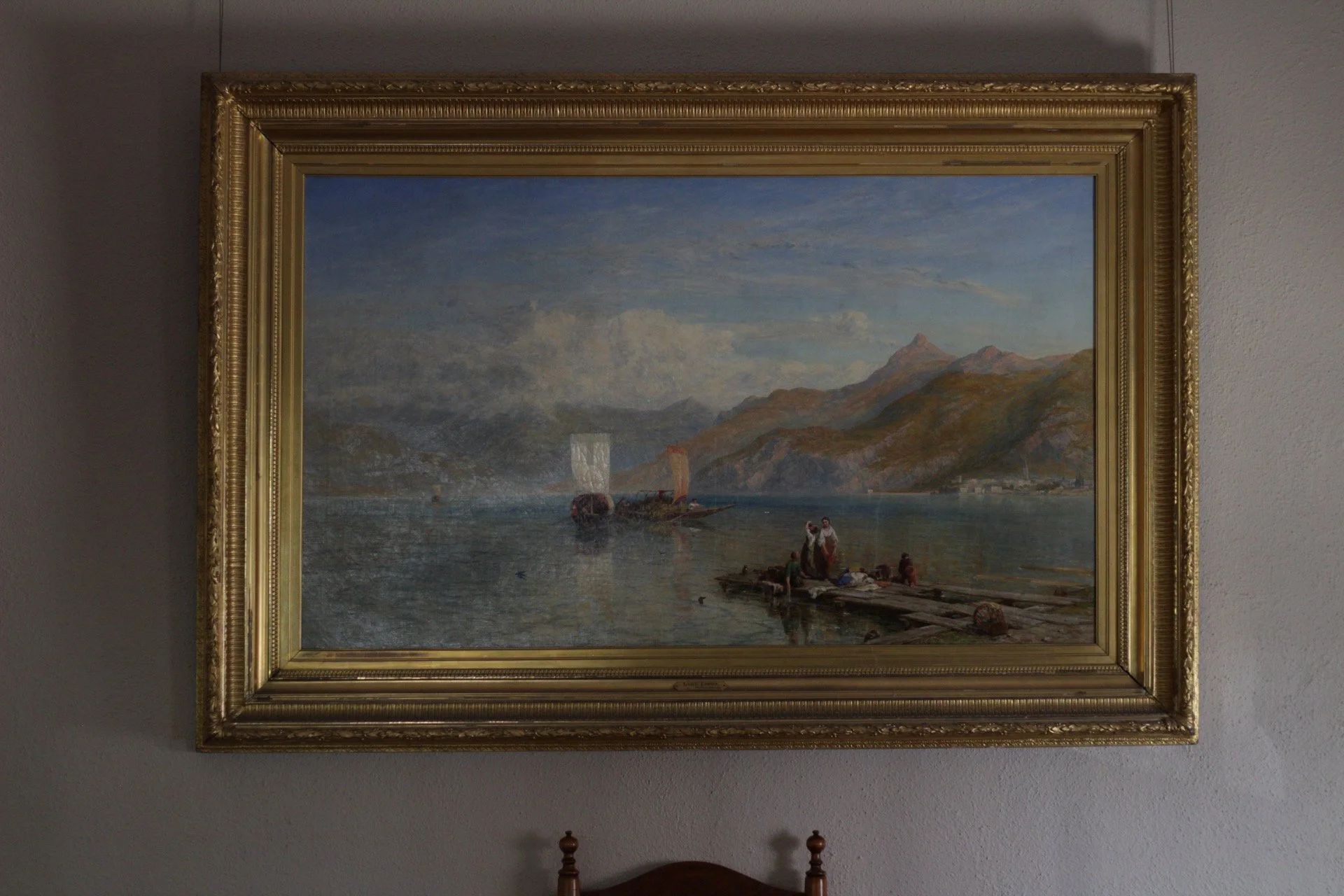 A framed landscape oil painting depicting a coastal scene with mountains in the background, a sailboat on the water, and people on the shoreline, hung on a gray wall.