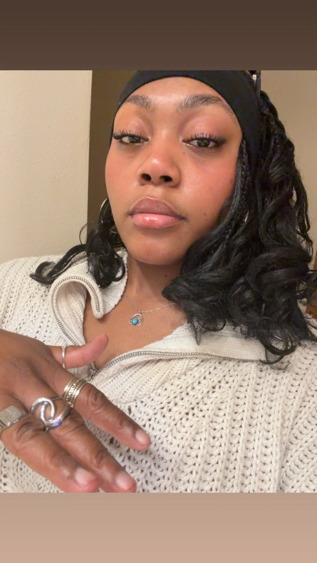 Close-up of a woman with dark curly hair wearing a beige knit sweater, multiple rings, and a necklace with a blue pendant, making a pointer gesture.