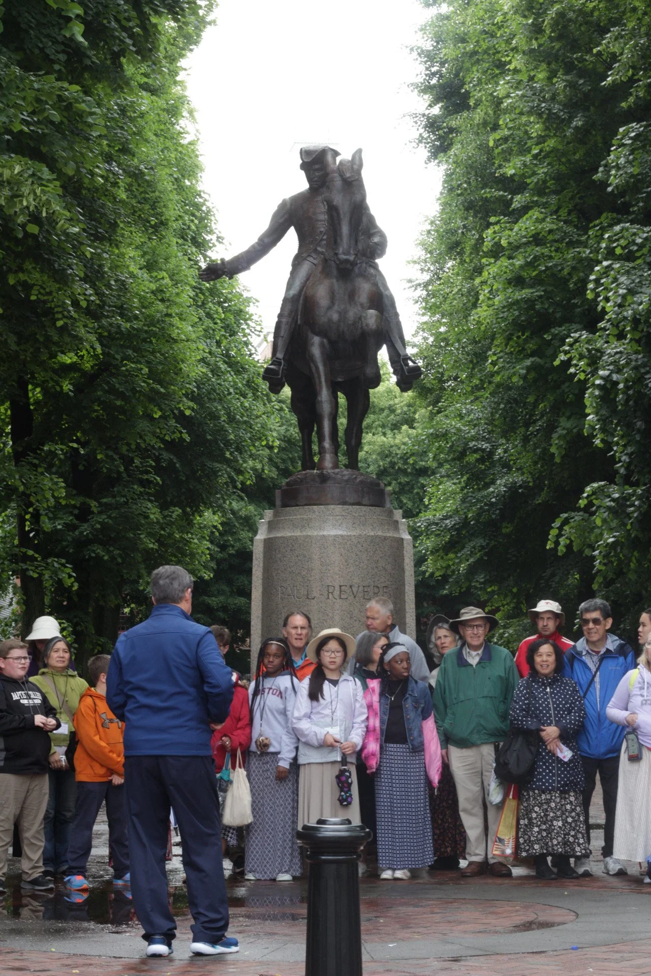 A group of diverse people gathered in front of a statue of Paul Revere, who is depicted riding a horse, in a park with lush green trees surrounding them.