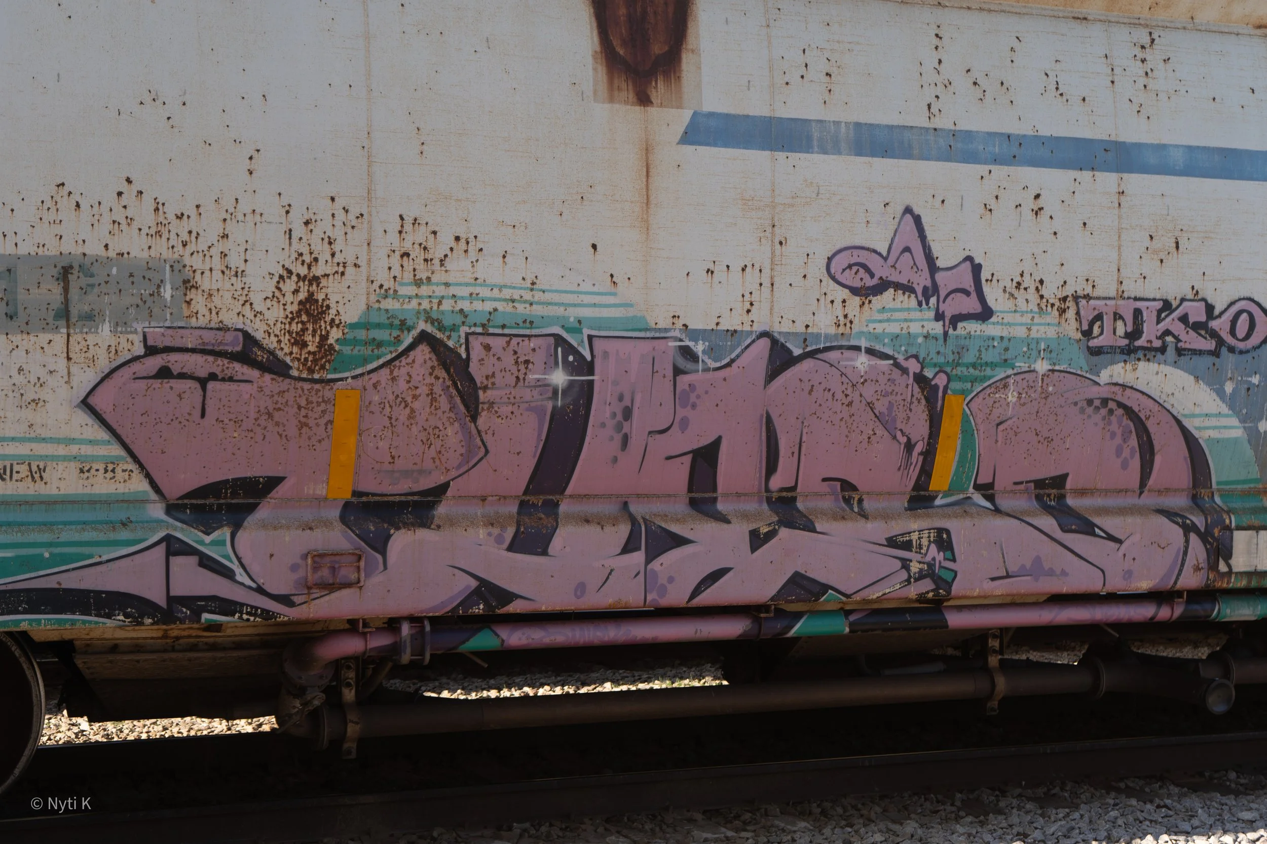Graffiti on a train car with rust spots and worn paint, featuring stylized letters in purple, teal, and black, with a small star-like detail. The image also shows the undercarriage and wheel assembly of the train.